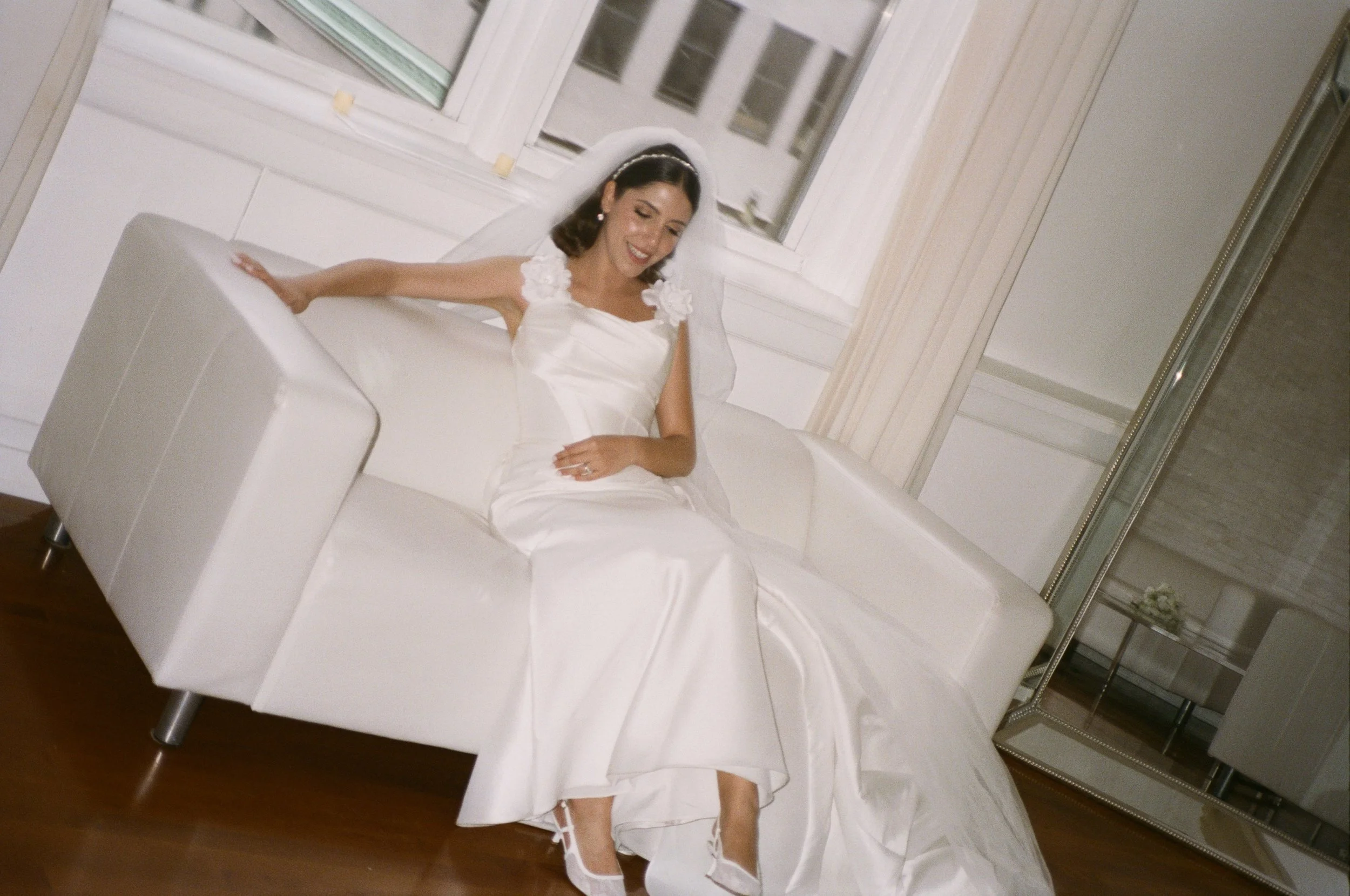 A bride sitting on a white couch in a room with large windows, dressed in a white wedding gown and veil, smiling and looking down.