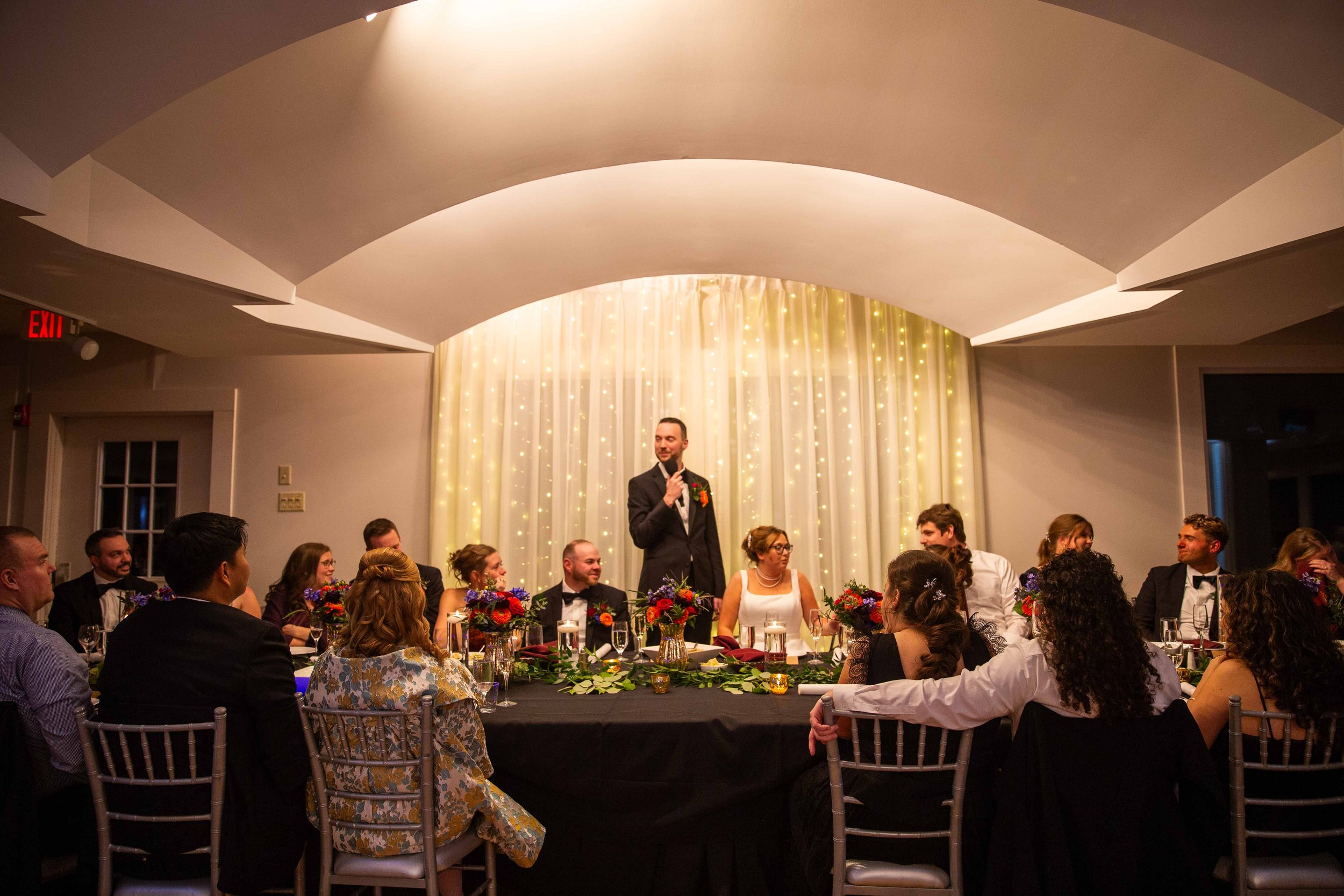 A wedding reception with a man giving a speech at a decorated head table, surrounded by seated guests at a formal event.