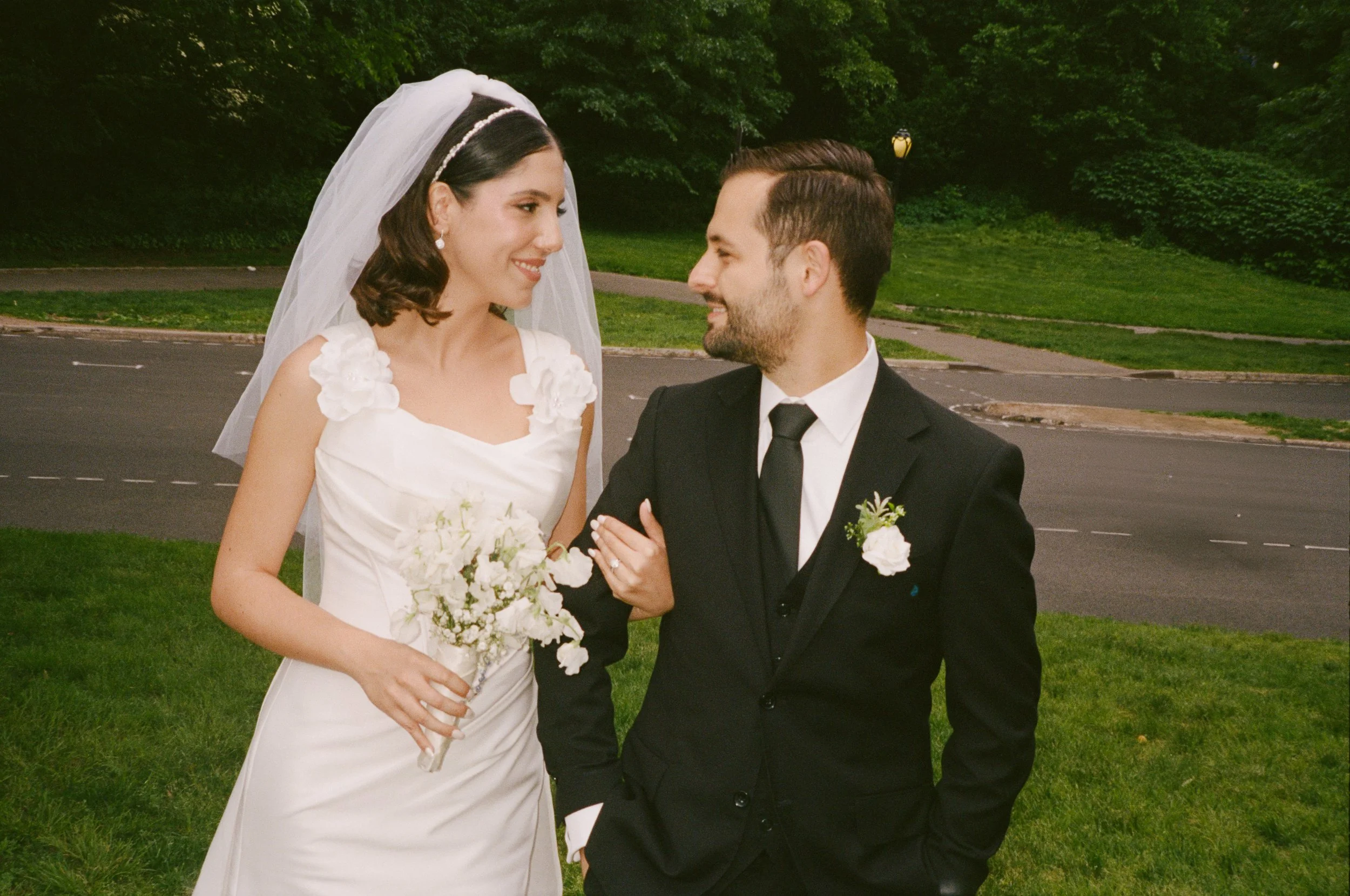 A bride and groom smiling at each other outdoors on a grassy area.