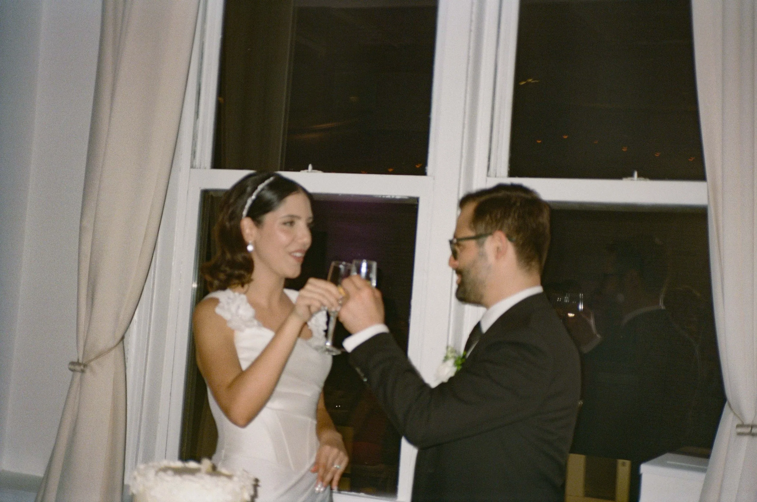 A bride and groom celebrating their wedding inside a room with large windows and curtains, raising glasses for a toast. The bride is in a white gown with floral details and the groom is in a black tuxedo.
