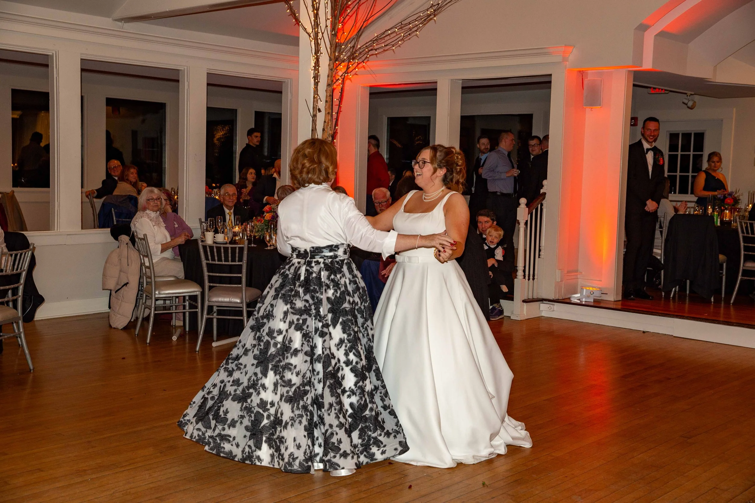 Two women dancing together at a wedding reception, with guests seated and standing in the background, decorated with warm lighting.