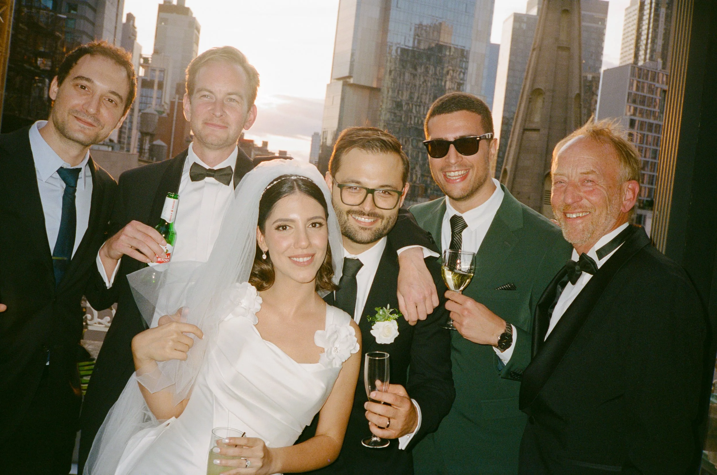 Group of six people celebrating at a wedding on a rooftop in a city, with a skyline of skyscrapers in the background, some holding drinks, friend group smiling and posing.