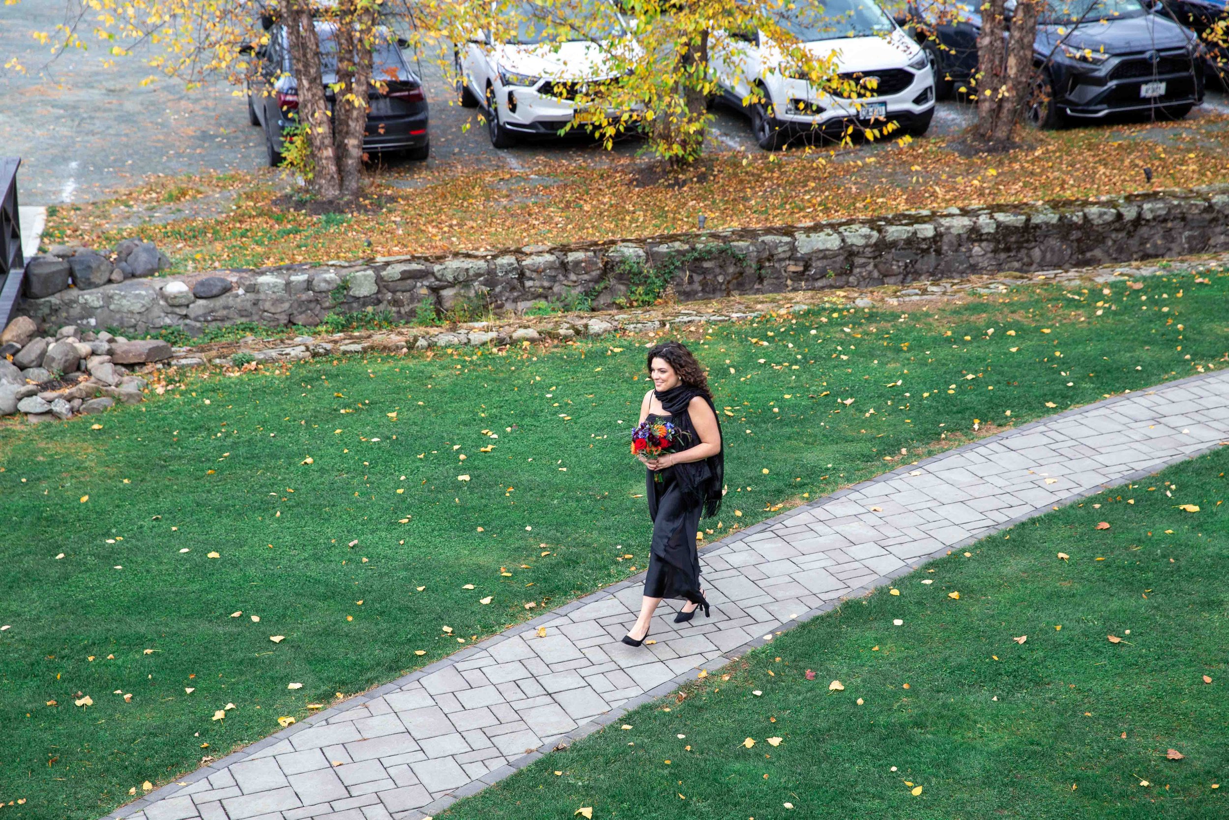 A woman walking on a curved stone pathway holding a bouquet of flowers, with green grass, fallen autumn leaves, trees, and parked cars in the background.
