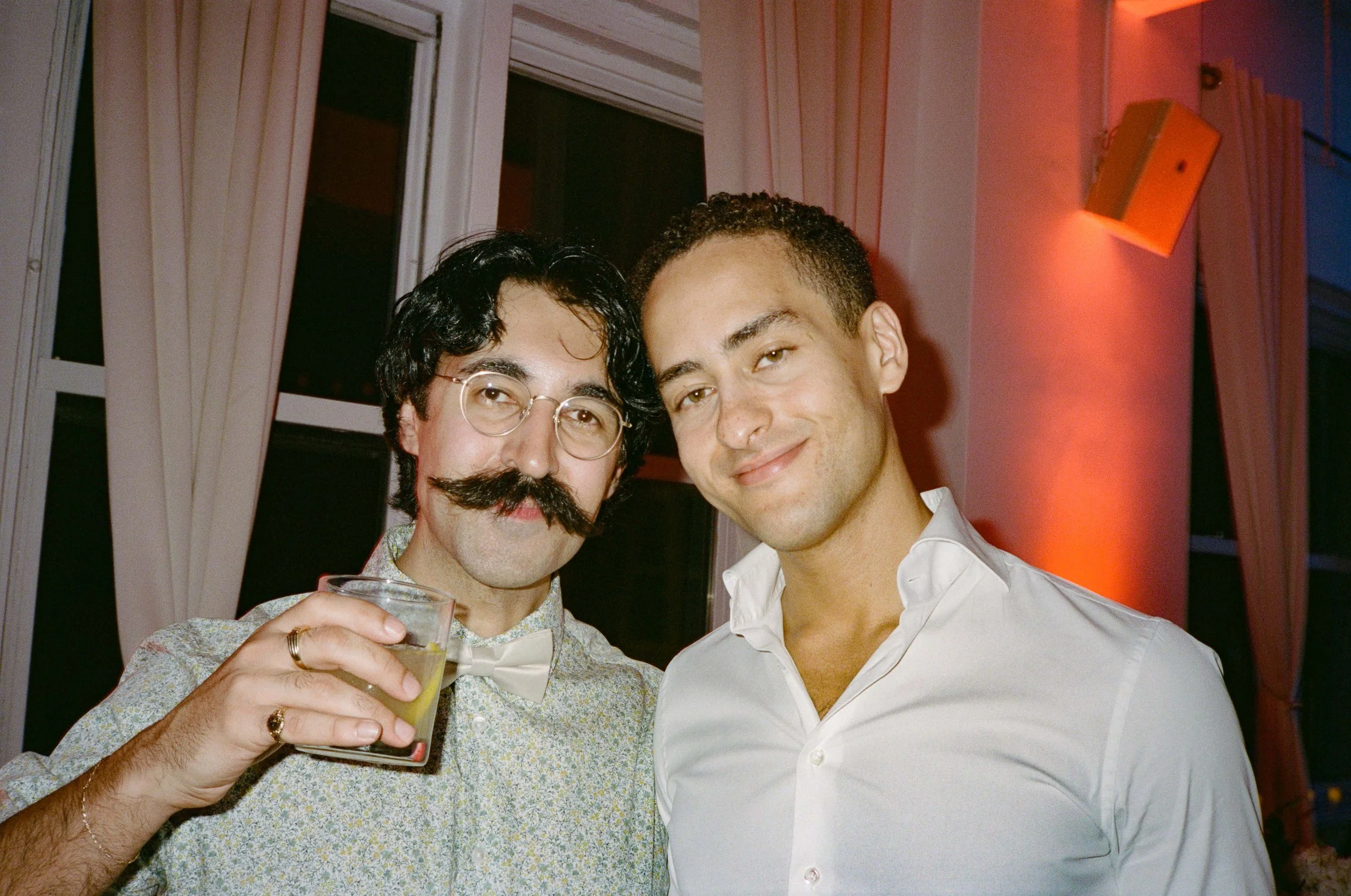 Two men smiling indoors, one with dark curly hair and glasses, the other with short dark hair, wearing a white shirt, in a room with pink curtains and an orange wall light.