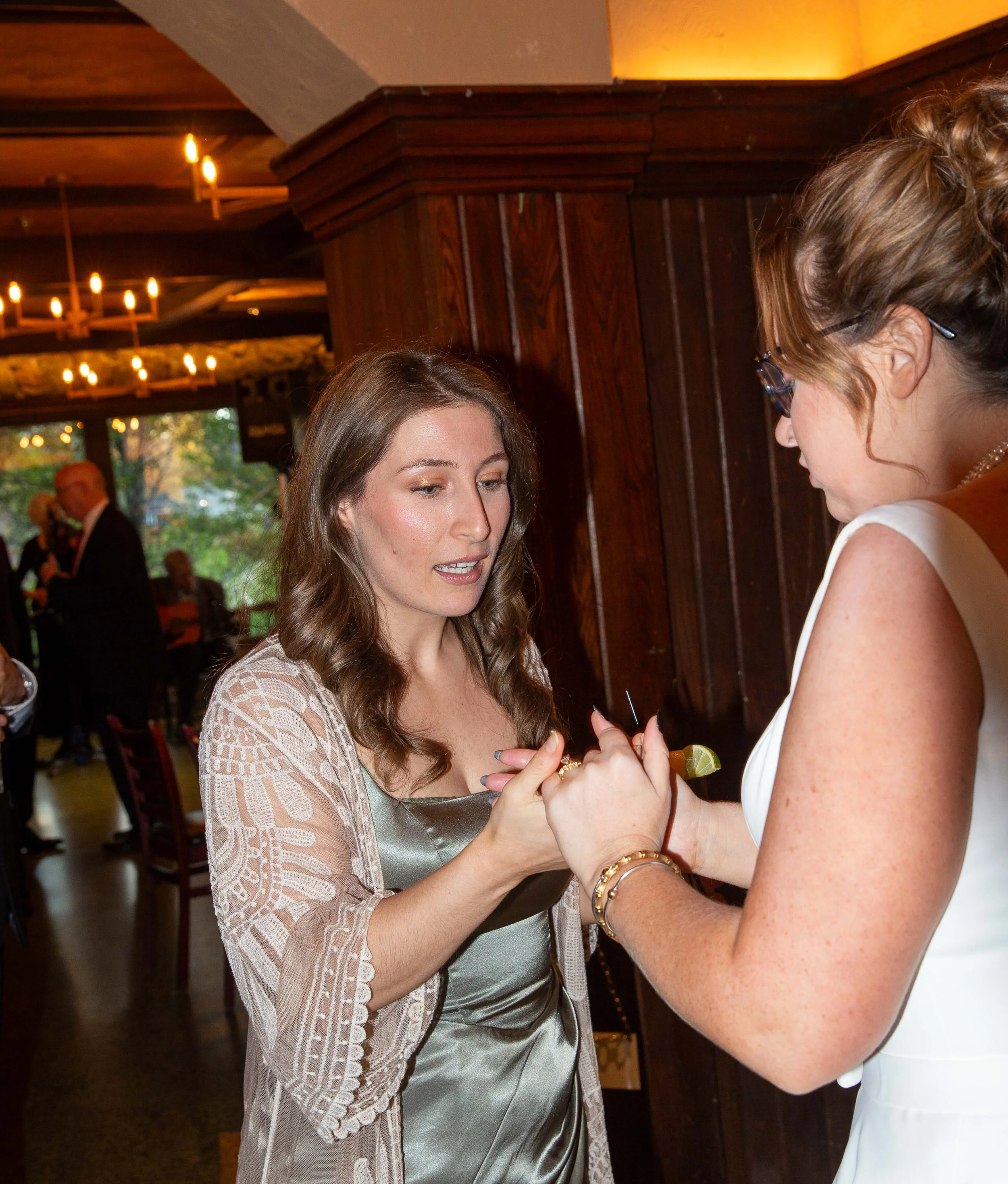 Two women are exchanging rings at a wedding ceremony inside a warmly lit venue with wooden walls and chandeliers. One woman is in a satin dress and the other in a white gown with glasses.