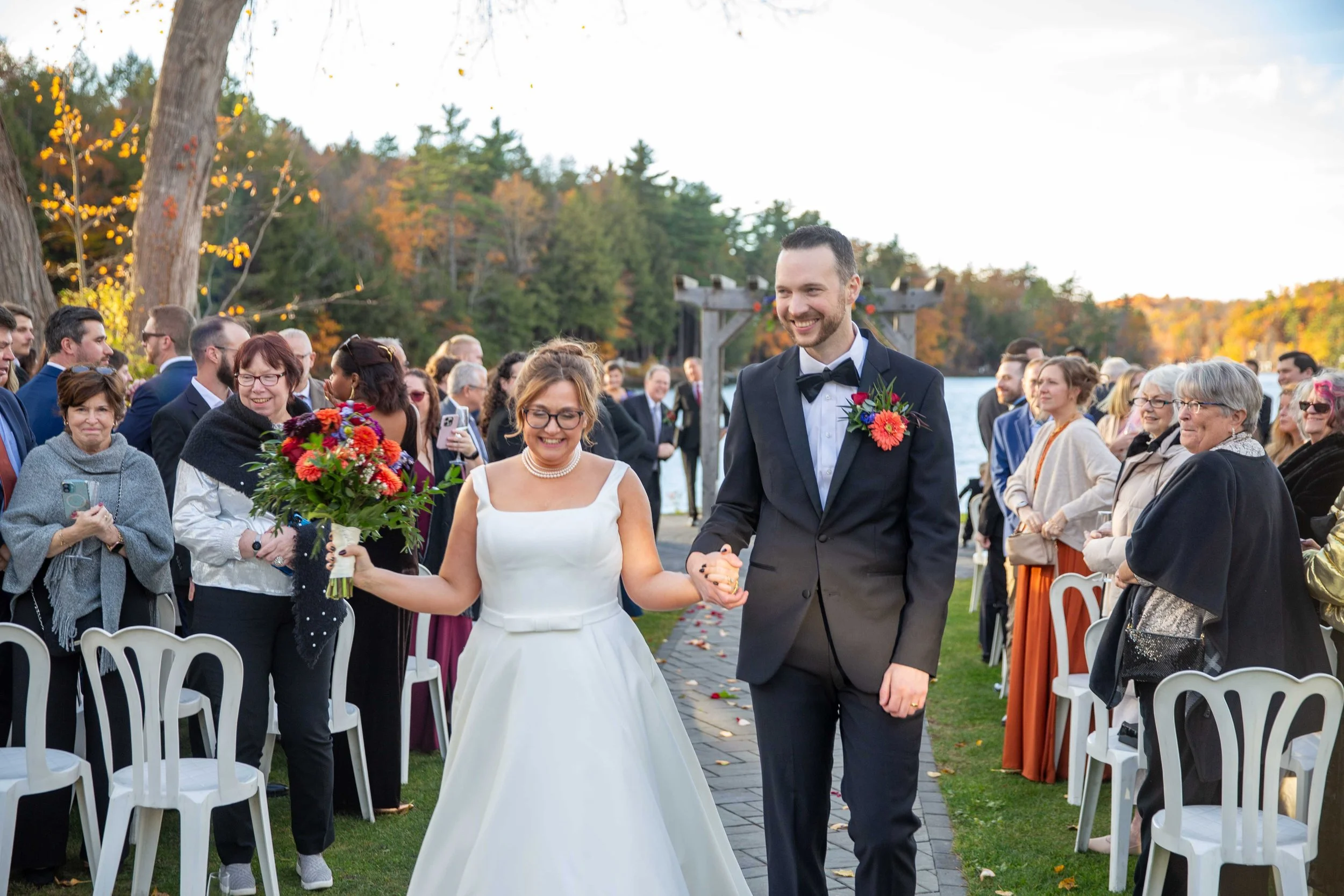 A bride and groom walking hand in hand down an aisle outdoors at sunset, surrounded by wedding guests, with trees and a lake in the background.