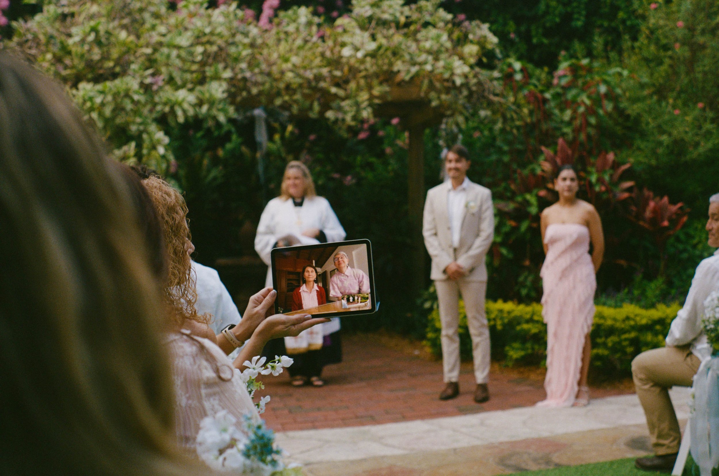 A couple dressed in wedding attire standing outdoors during a wedding ceremony, with a woman taking a photo on a tablet. There are guests, greenery, and flowers in the background.
