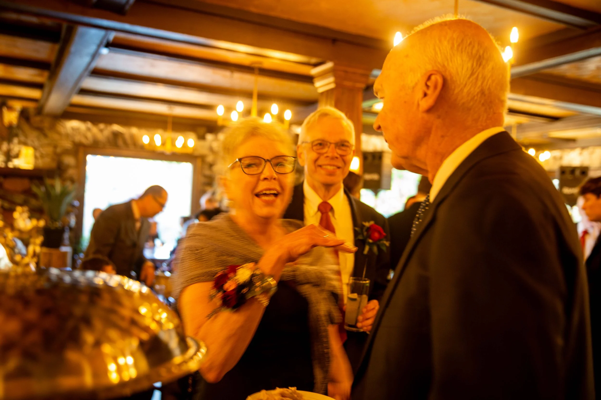 Three elderly people at a social gathering, smiling and talking to each other inside a warmly lit room with wooden beams, chandeliers, and stone walls.