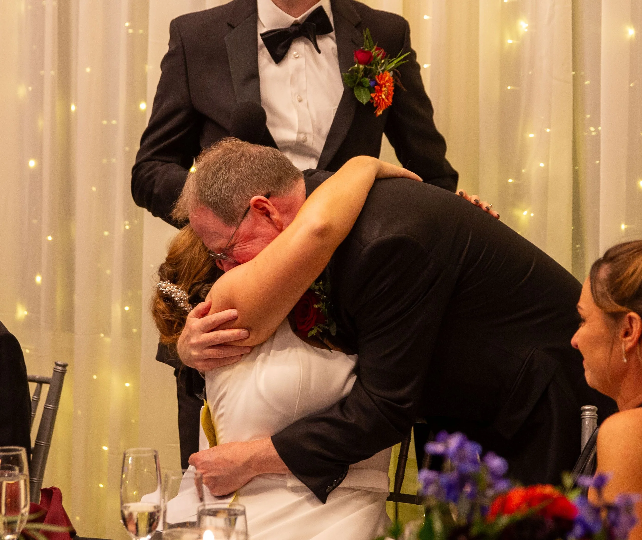 A man in a tuxedo kisses a woman in a white dress on the cheek at a wedding reception, with a man and woman smiling and sitting at a table nearby.