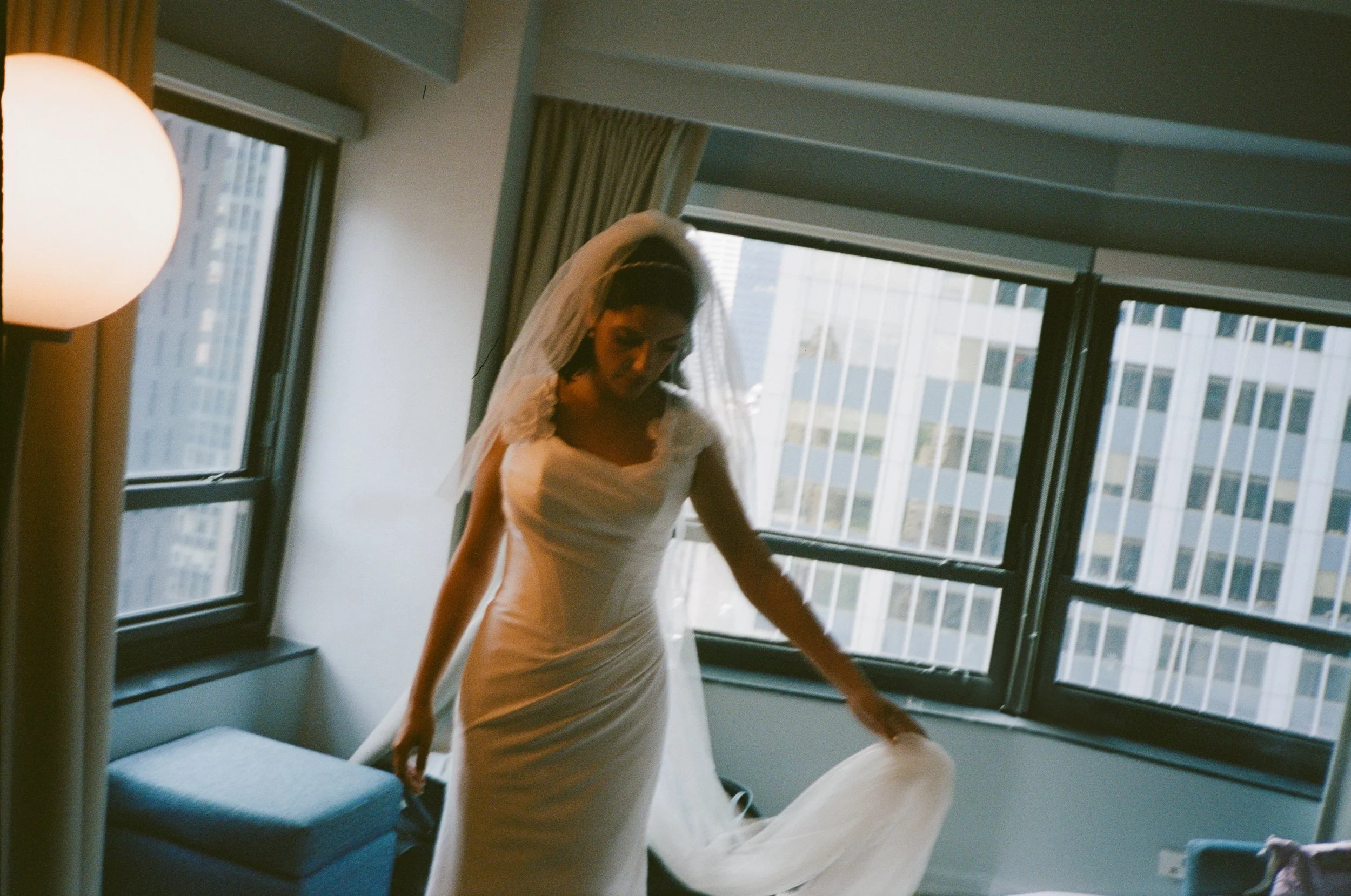 A bride in a white wedding dress and veil standing in a modern room with large windows and city view, adjusting her gown.