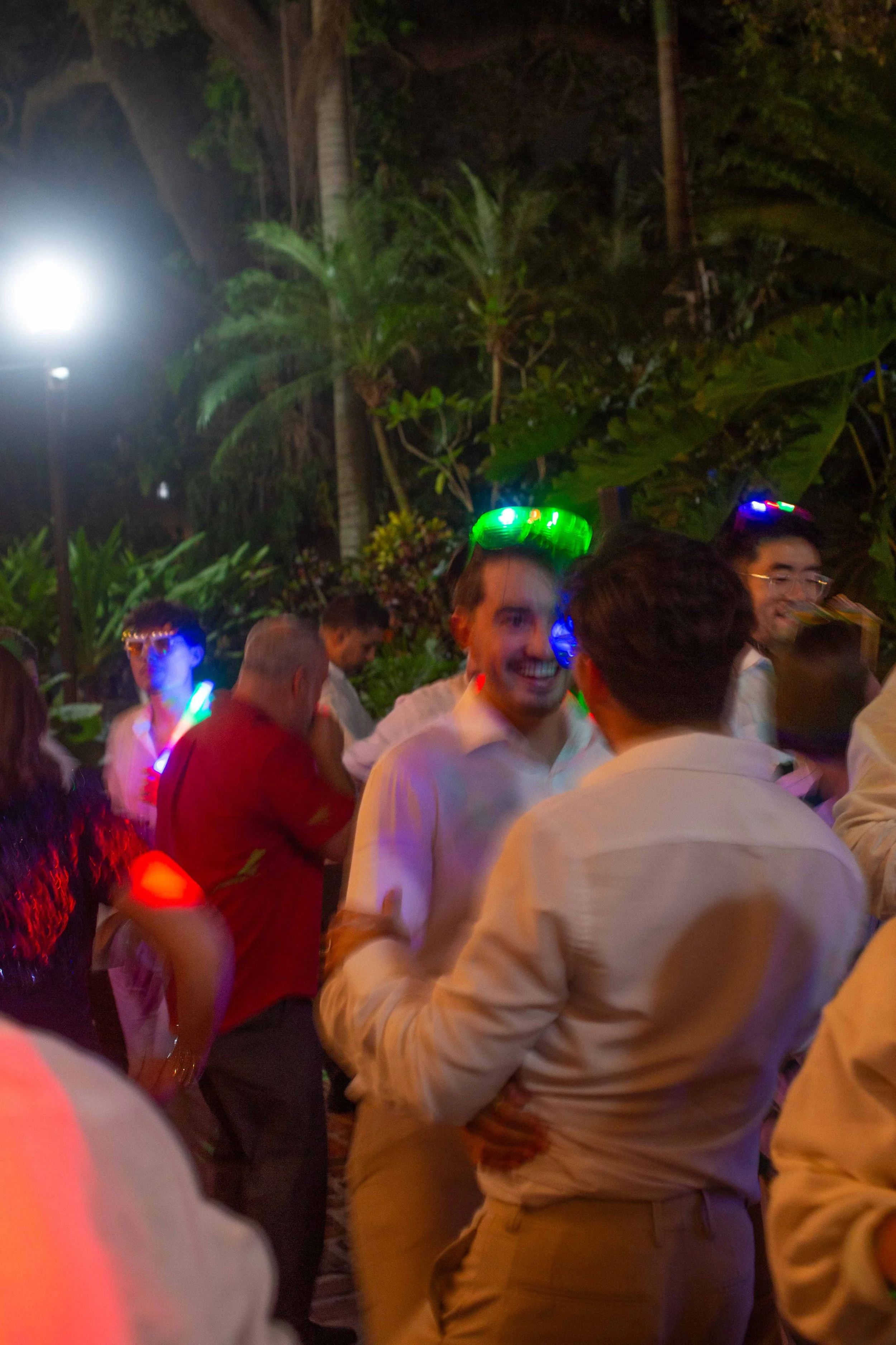 People dancing and socializing outdoors at night, with colorful lights and tropical plants in the background.