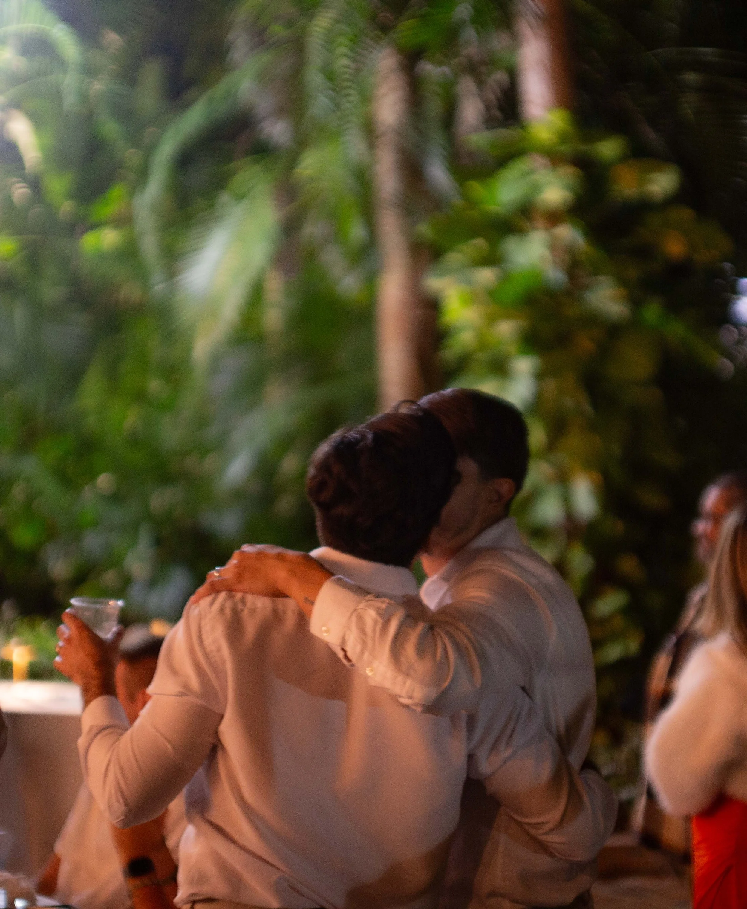 Two people embracing and kissing at an outdoor celebration, with others in the background and lush greenery around.