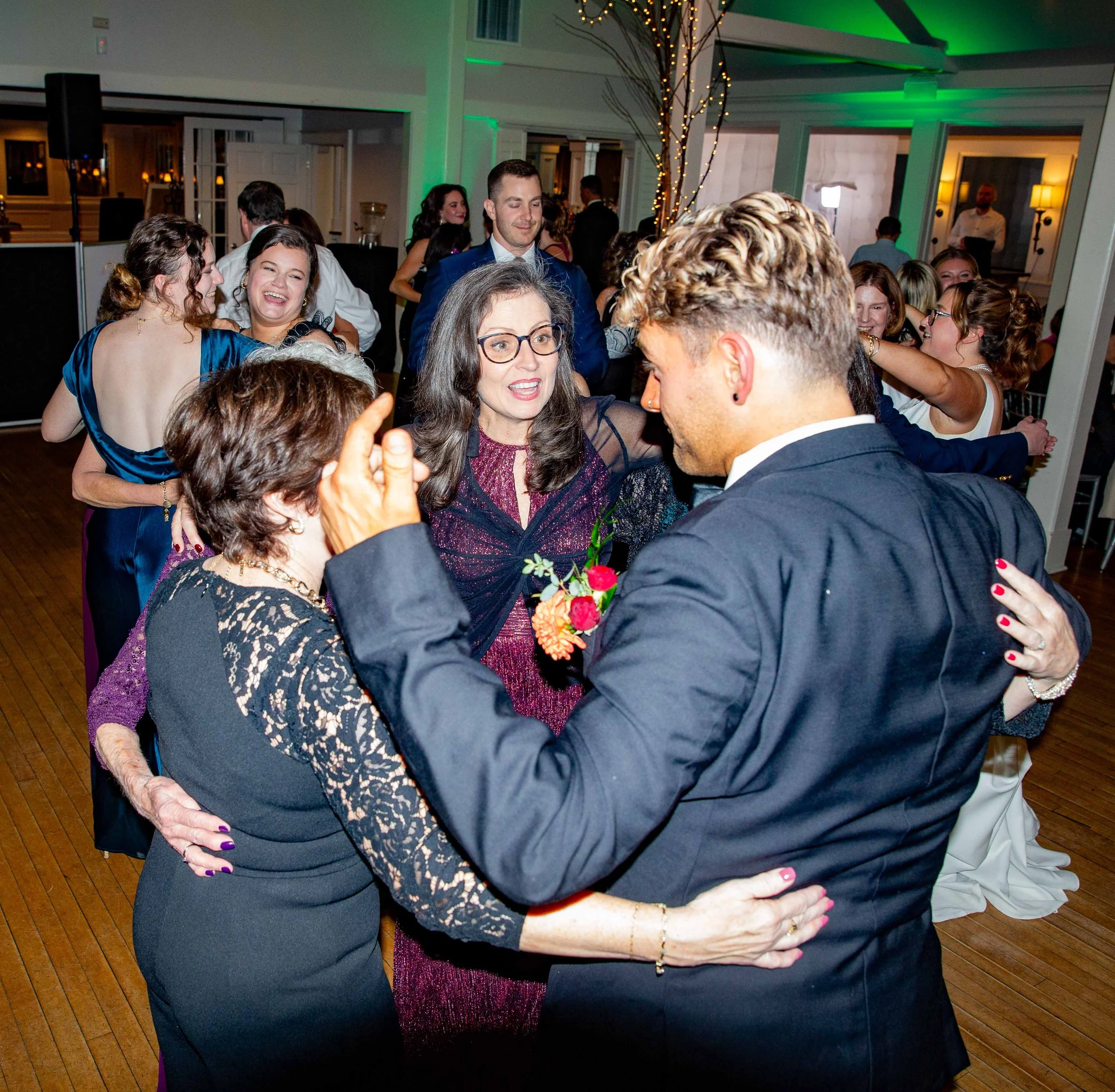 People dancing and celebrating at a wedding reception or party indoors, with string lights and a decorated tree in the background.