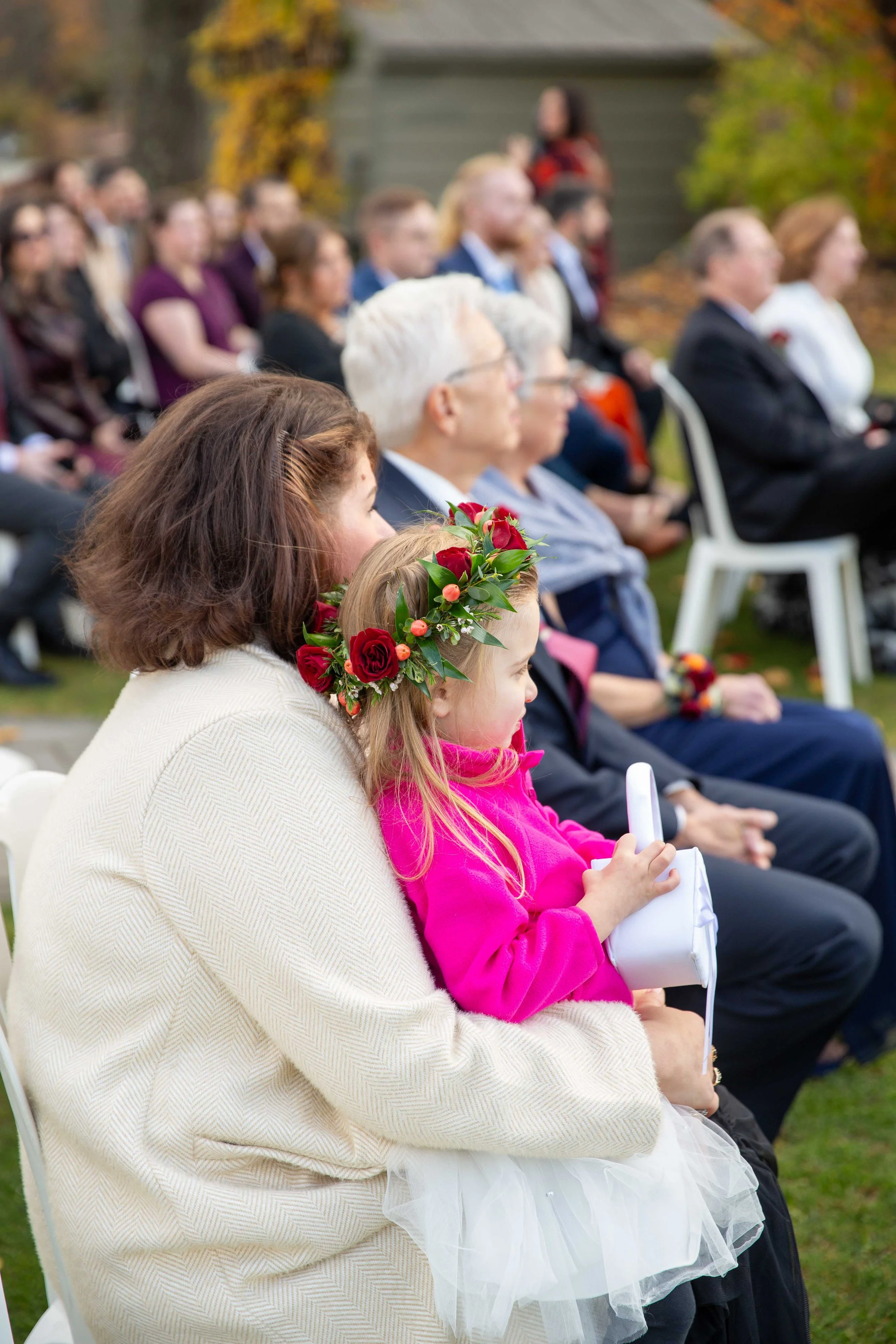 A woman with brown hair and a young girl wearing a pink jacket and a floral headband sitting outdoors at a wedding ceremony, surrounded by other seated guests.