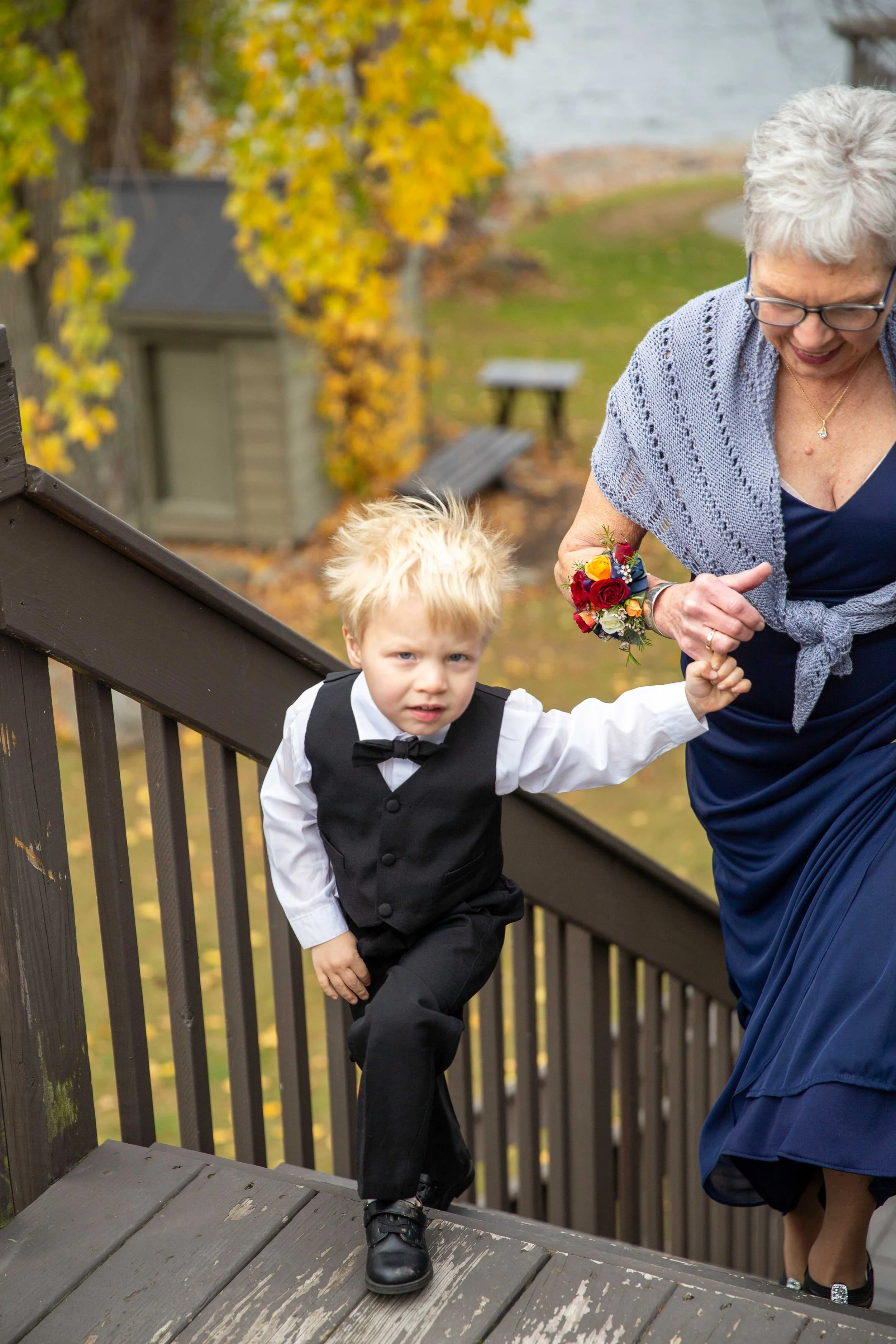 An elderly woman helping a young boy dressed in a formal suit and bow tie up a wooden staircase outdoors during autumn, with trees and a lake in the background.