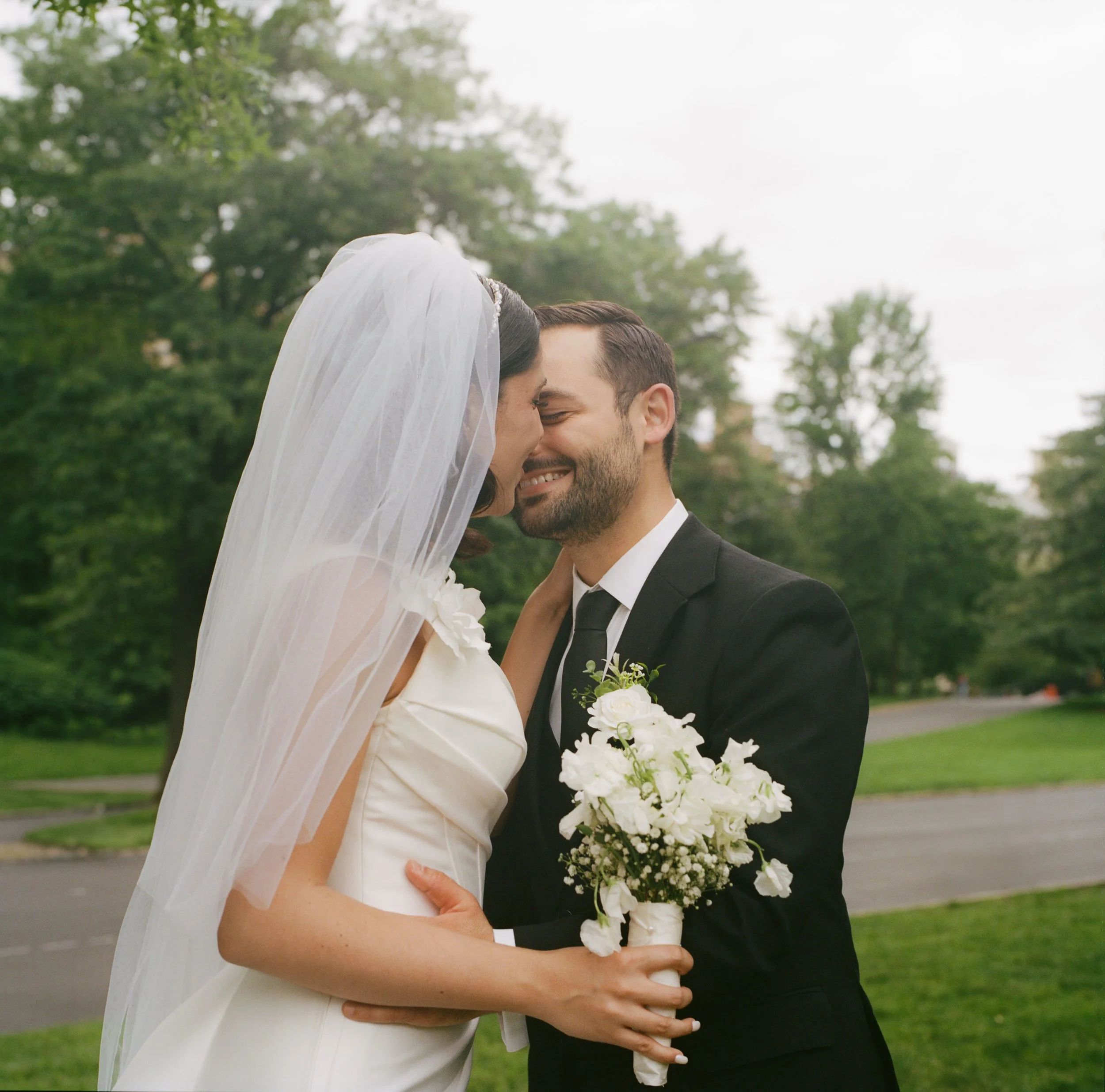 A bride and groom share a close moment outdoors, with the bride holding a white bouquet of flowers. The bride is wearing a white wedding dress and veil, and the groom is dressed in a black suit and tie. Green trees and an overcast sky are in the back