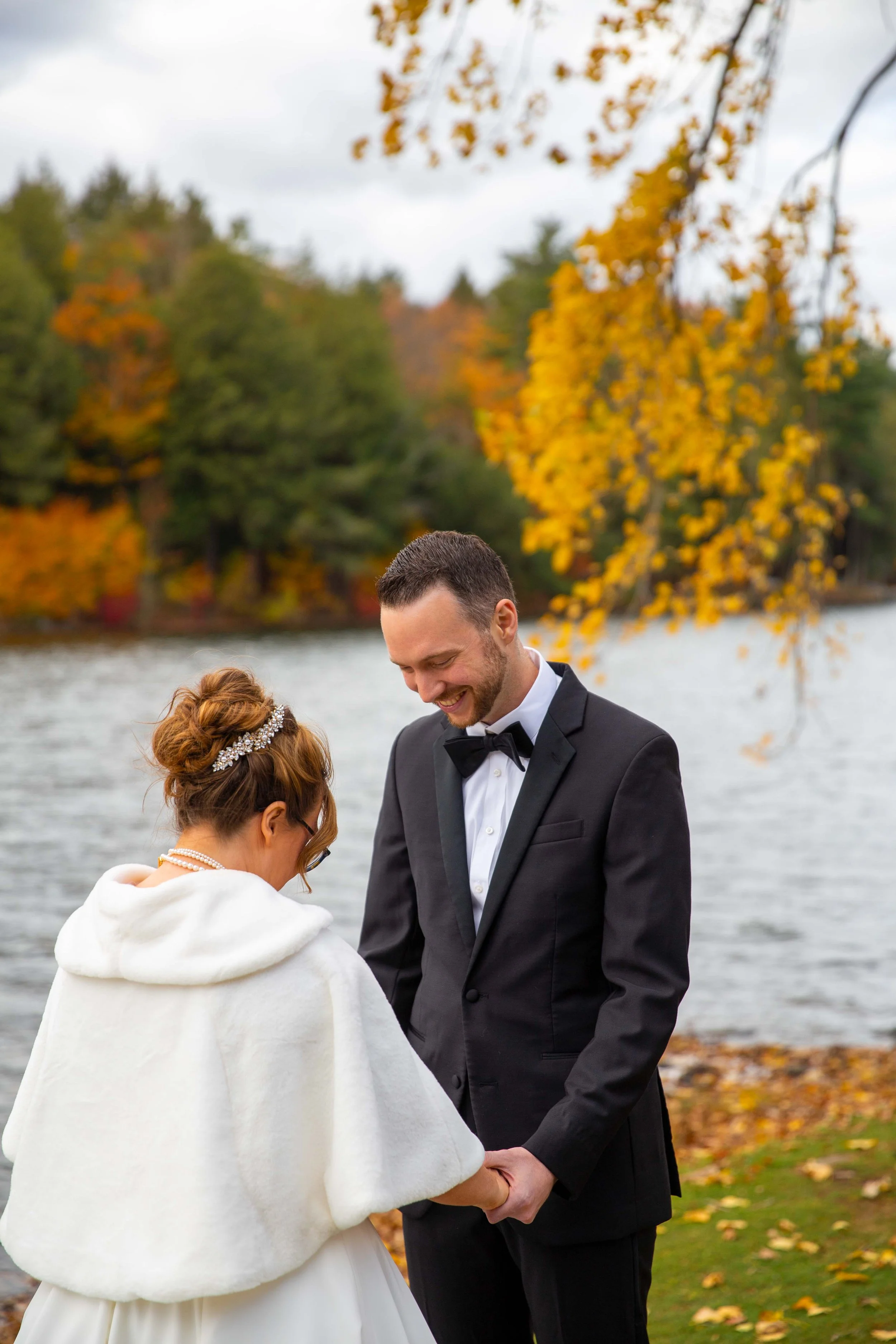 A bride and groom holding hands and smiling by a lake during fall with colorful autumn trees in the background.