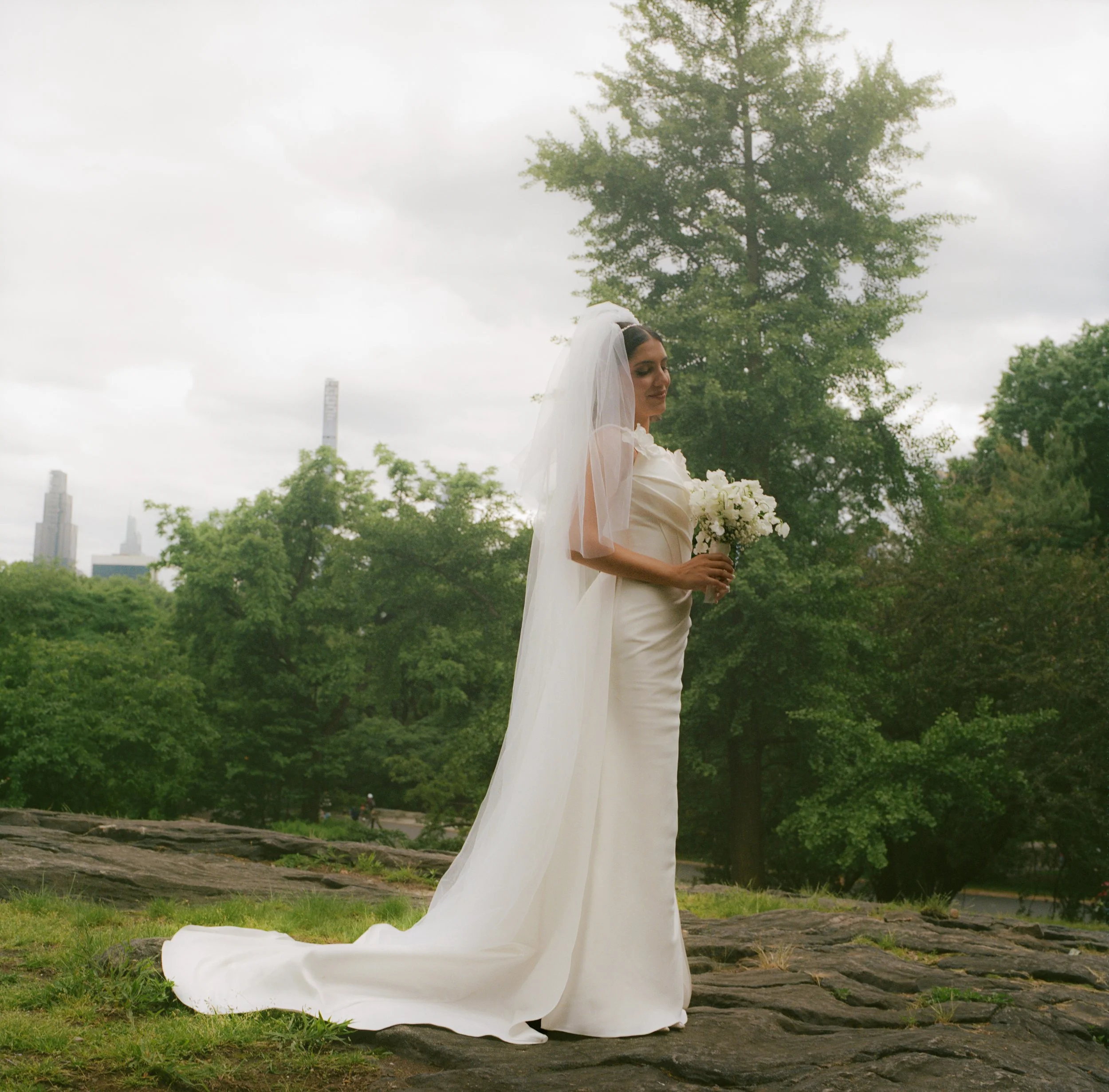 A bride in a white wedding gown with a long train and veil, holding a bouquet of white flowers, stands outdoors on a rocky surface with green trees and a city skyline in the background.