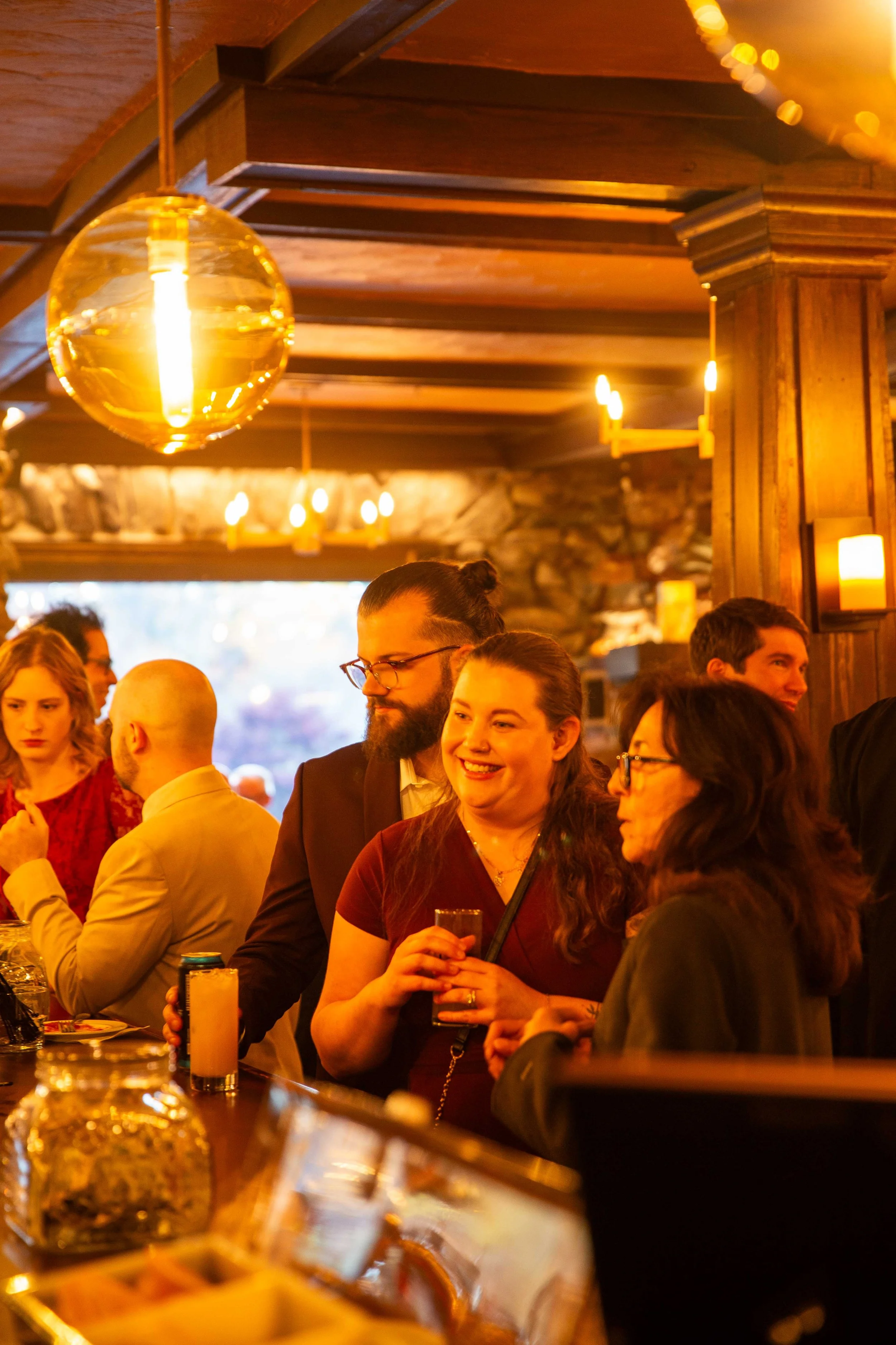 People socializing in a warmly lit bar or restaurant with wooden interior decor and hanging pendant lights.