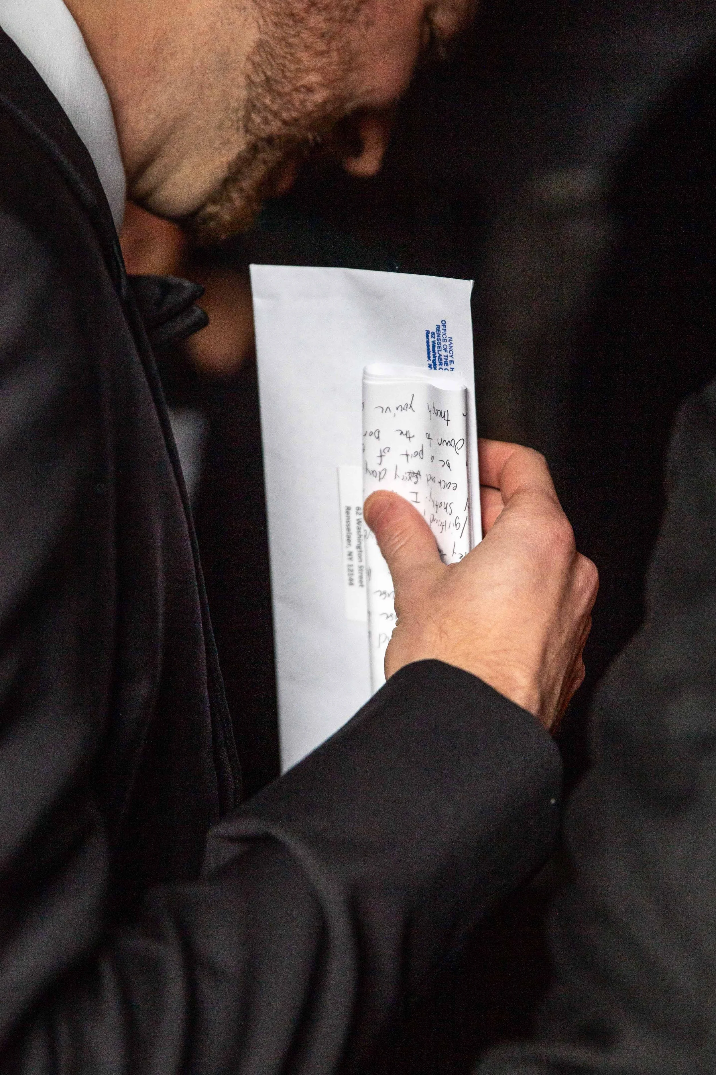 Close-up of a person holding an envelope and a handwritten note inside a tube, with part of a face visible, in a dark setting.
