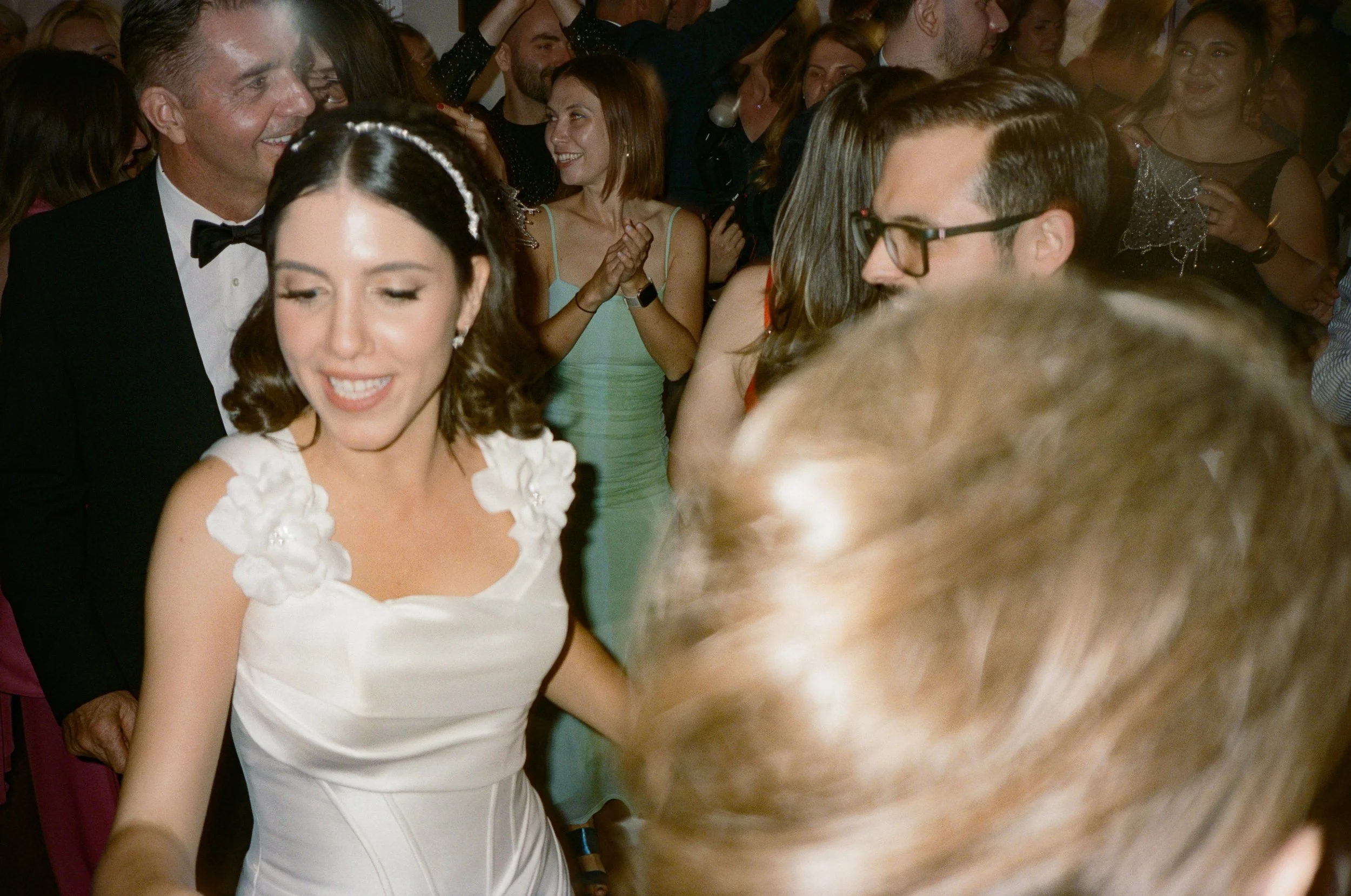People dancing at a wedding reception, with the bride in a white dress in the foreground, smiling, surrounded by guests in formal attire.