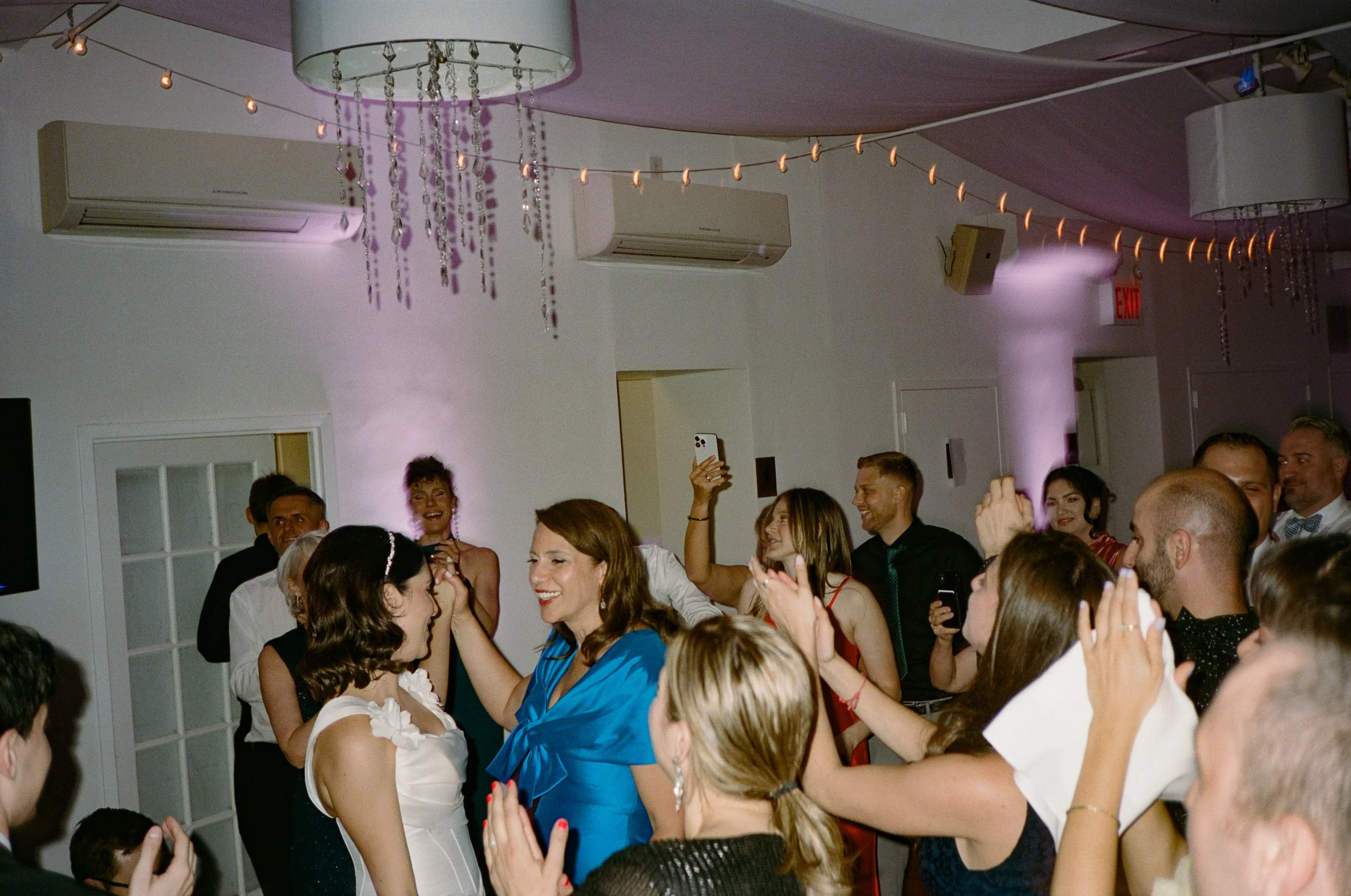 Group of people at a wedding celebration, smiling, dancing, and taking photos indoors with string lights and chandeliers.