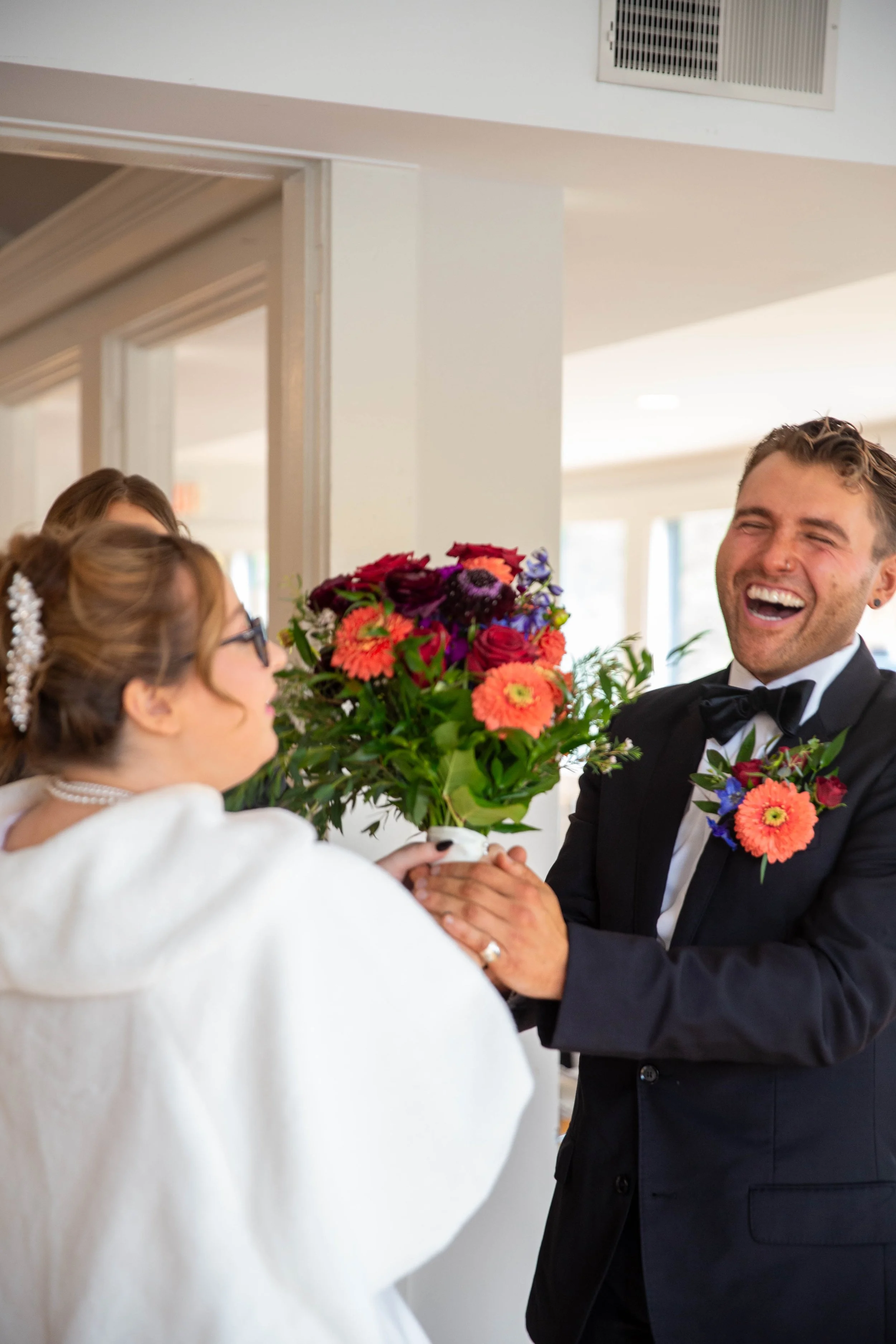 A groom in a tuxedo with a boutonniere smiling and holding a bouquet of colorful flowers, presenting it to a bride dressed in white with glasses, as they share a joyful moment during a wedding celebration indoors.
