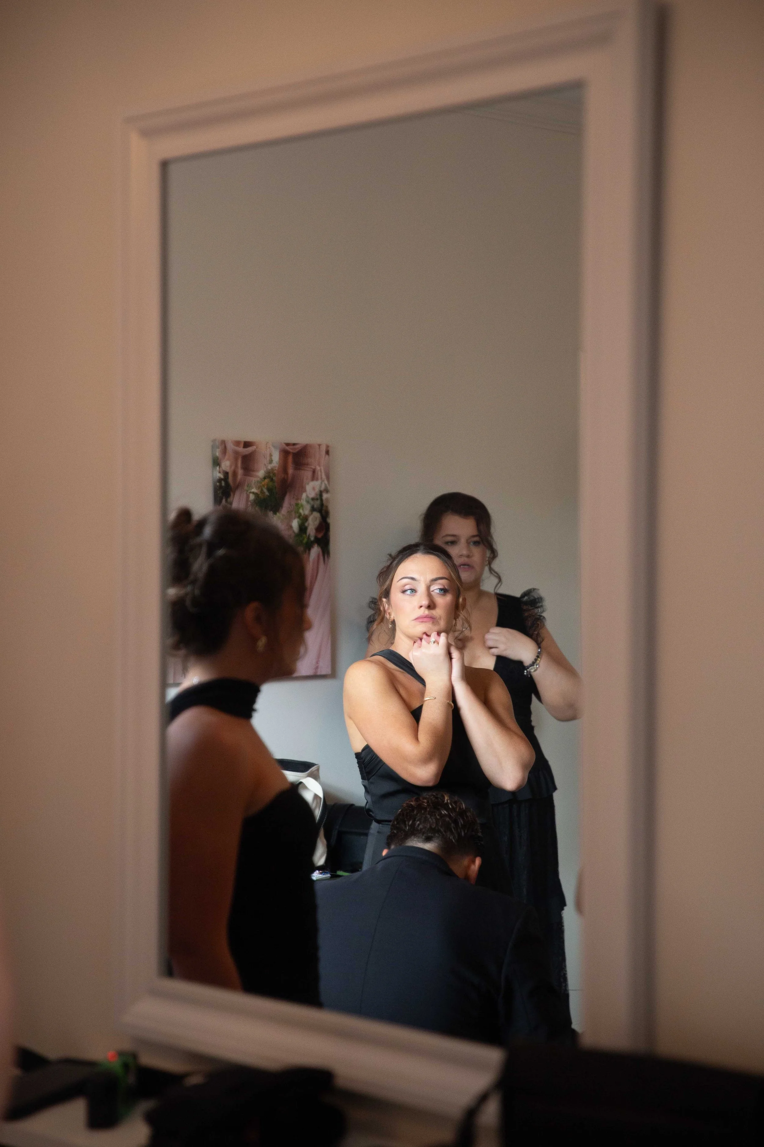 Reflection in a mirror shows three women preparing for an event, with one woman adjusting her hair, another with hands near her collar, and a third woman in the background.