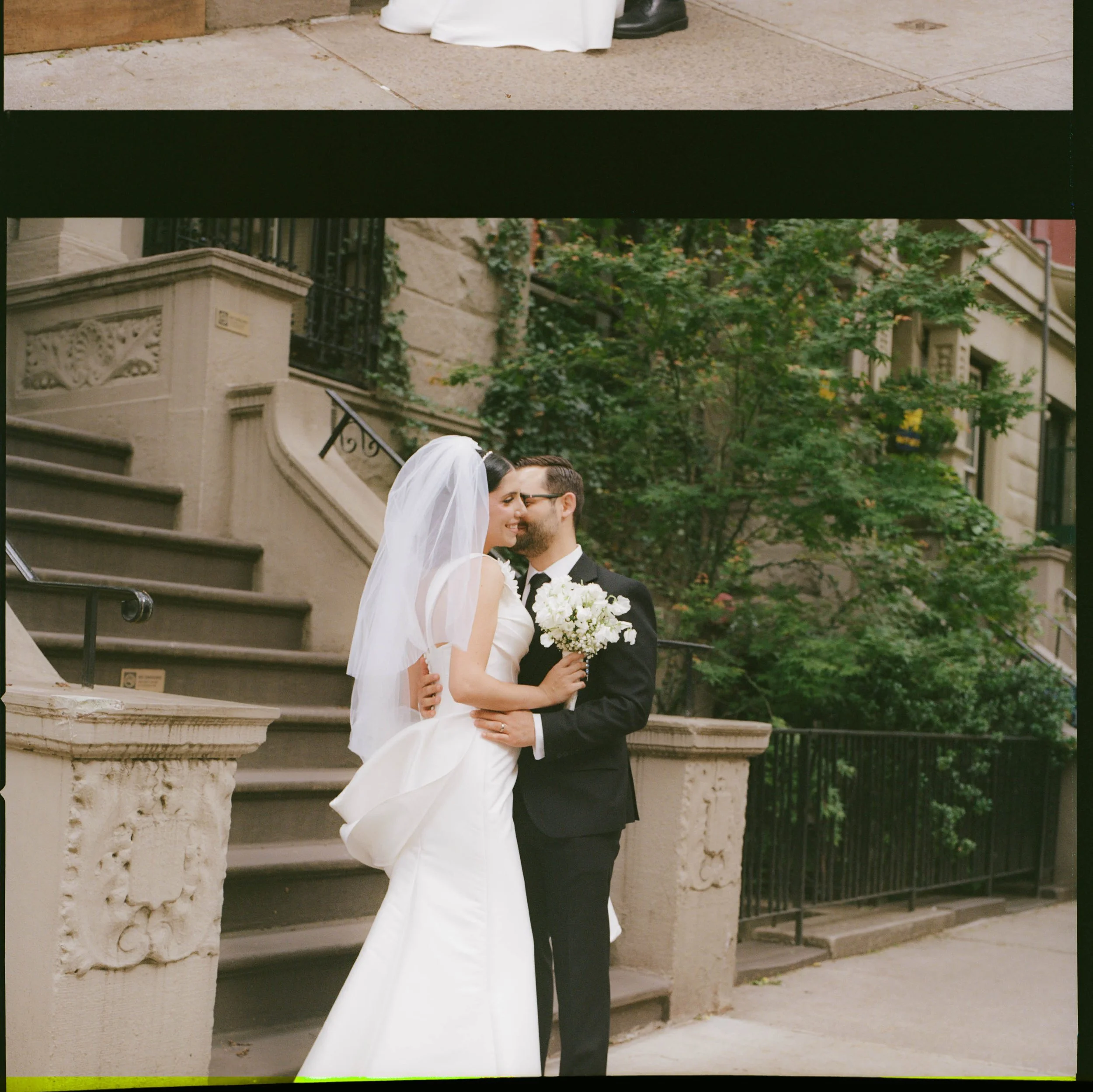 A bride and groom standing close on a city sidewalk with stone stairs and green trees in the background. The bride is holding a bouquet, and their faces are close as they smile.