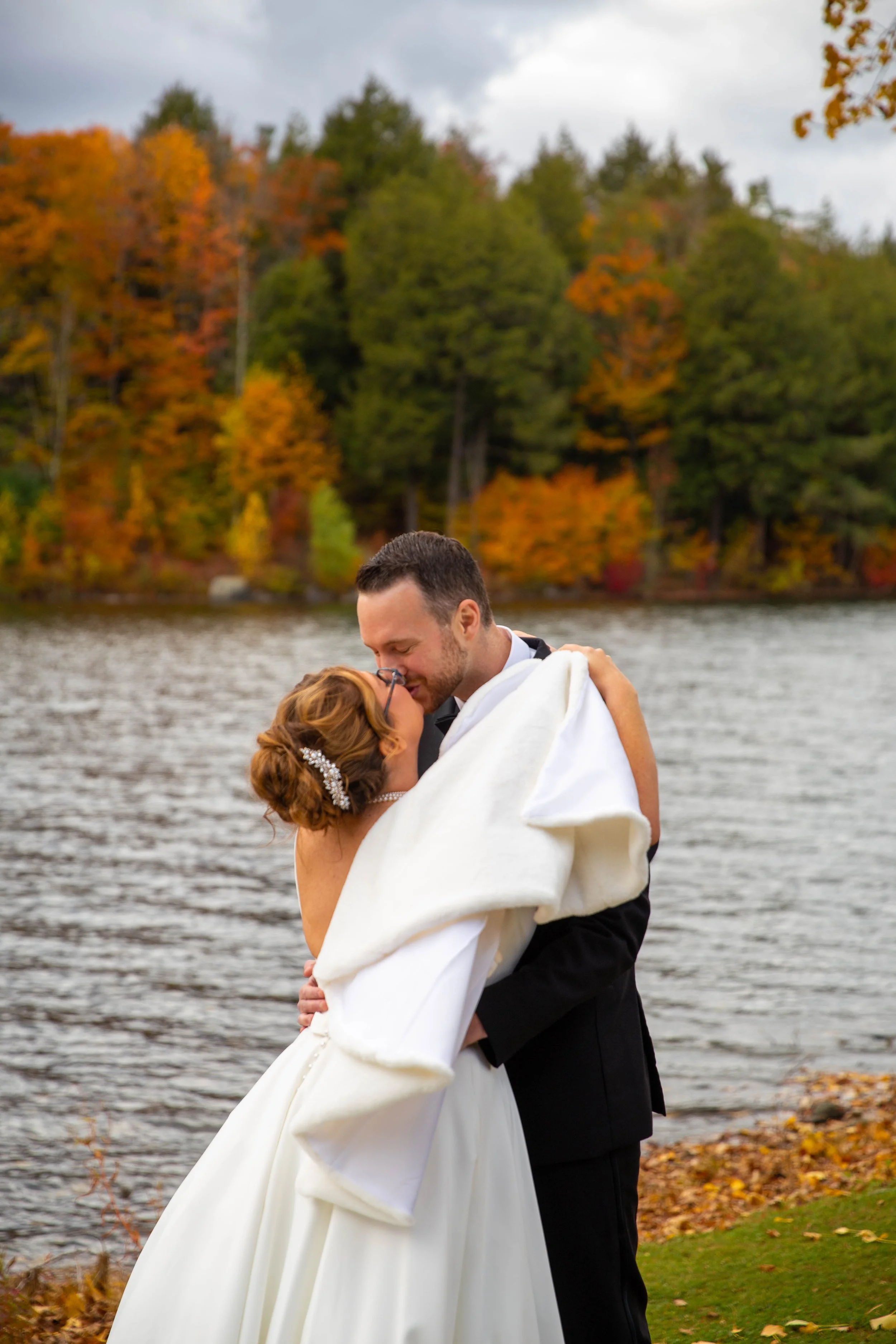 A bride and groom sharing a kiss outdoors by a lake with autumn trees in the background.