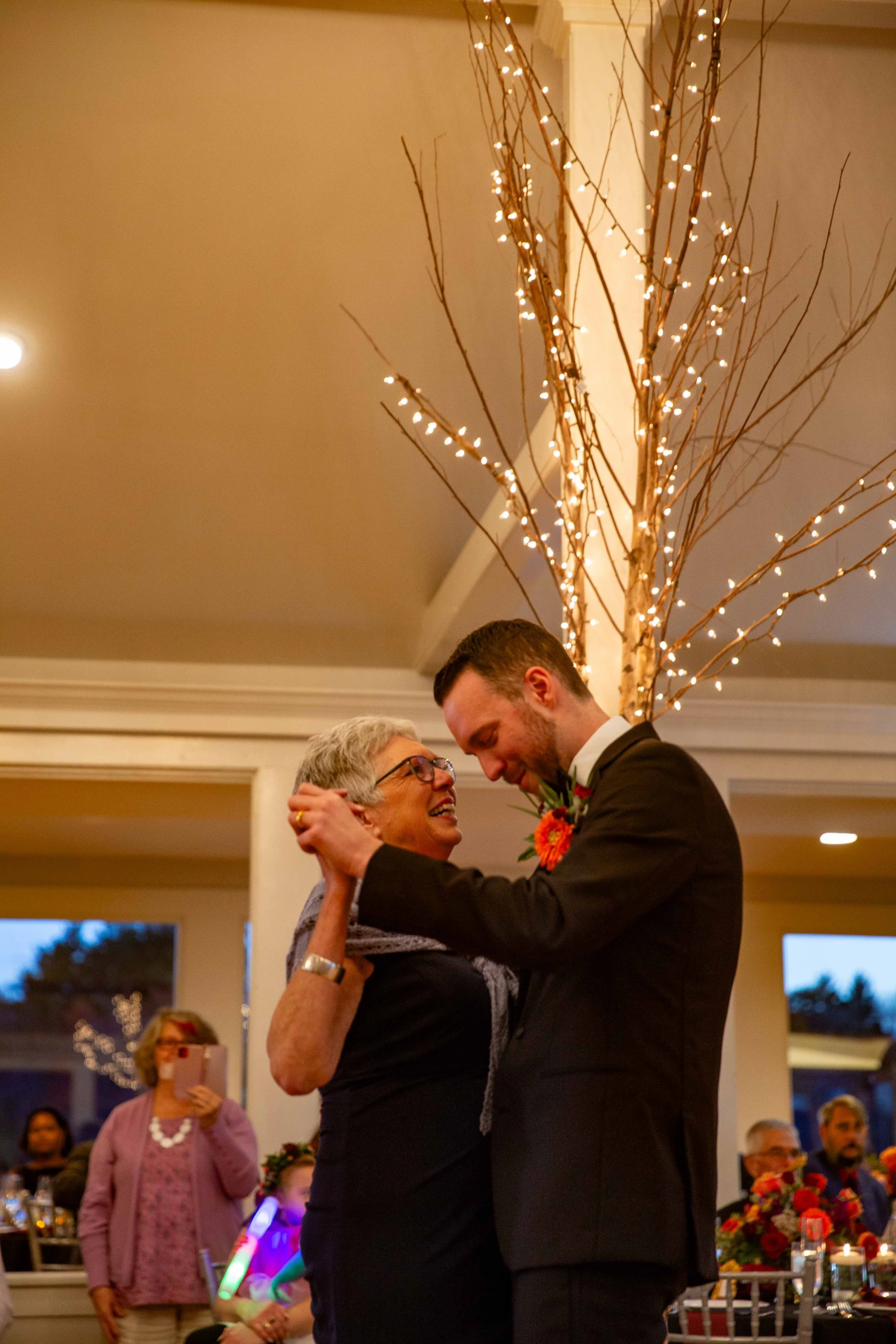 A young man in a black suit shares a dance with an older woman in a black outfit at a celebration, with festive lights and other guests watching, indoors.