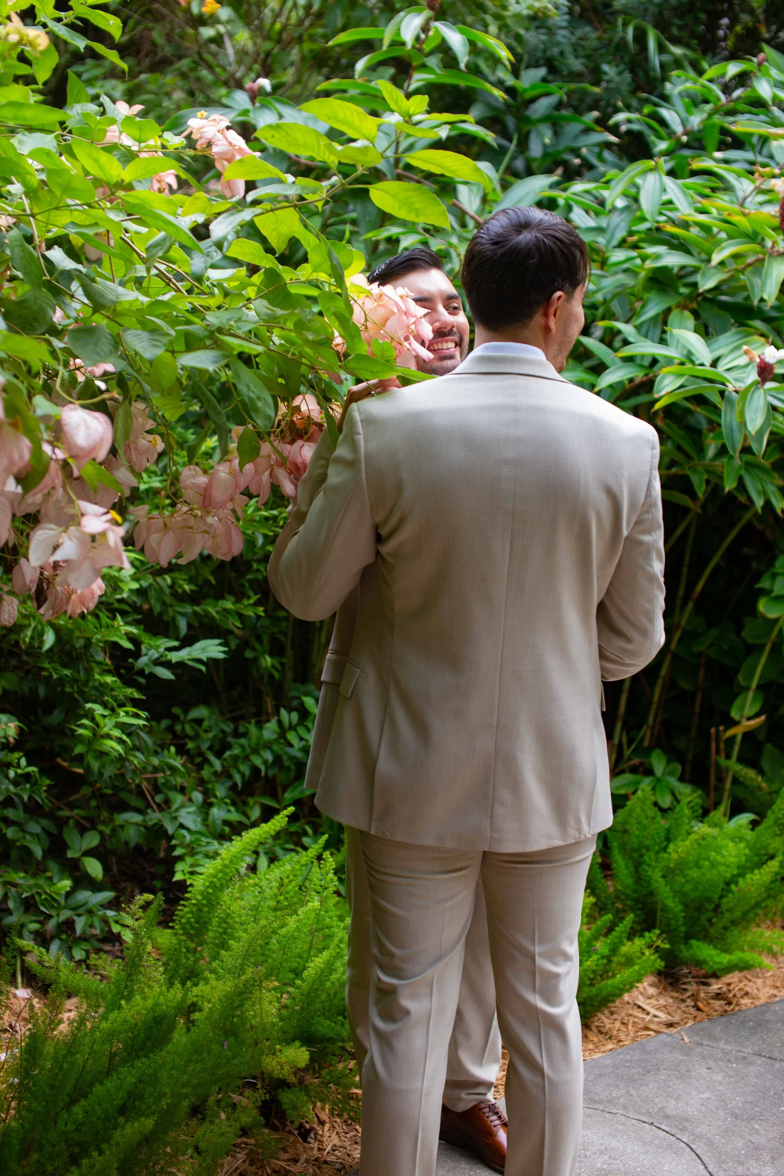 Two men in beige suits standing close to one another among lush green and pink flowering plants, smiling and engaging in a conversation in a garden setting.