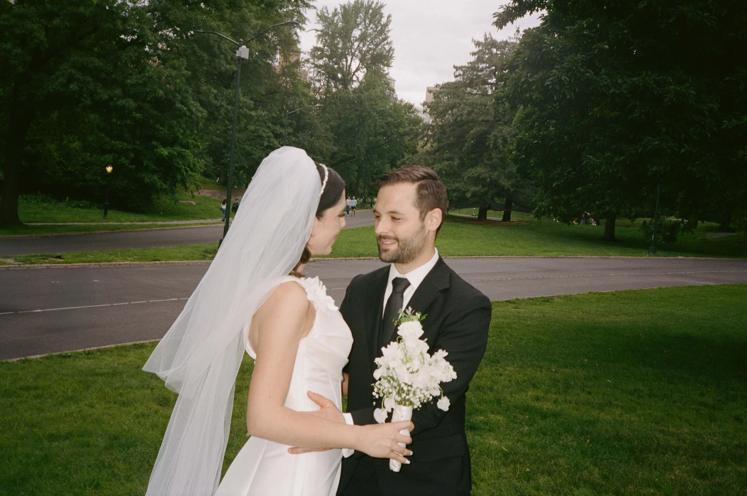A bride and groom stand close together in a park, looking into each other's eyes. The bride wears a white wedding dress with a veil, and the groom wears a black suit with a tie. The groom holds a bouquet of white flowers.