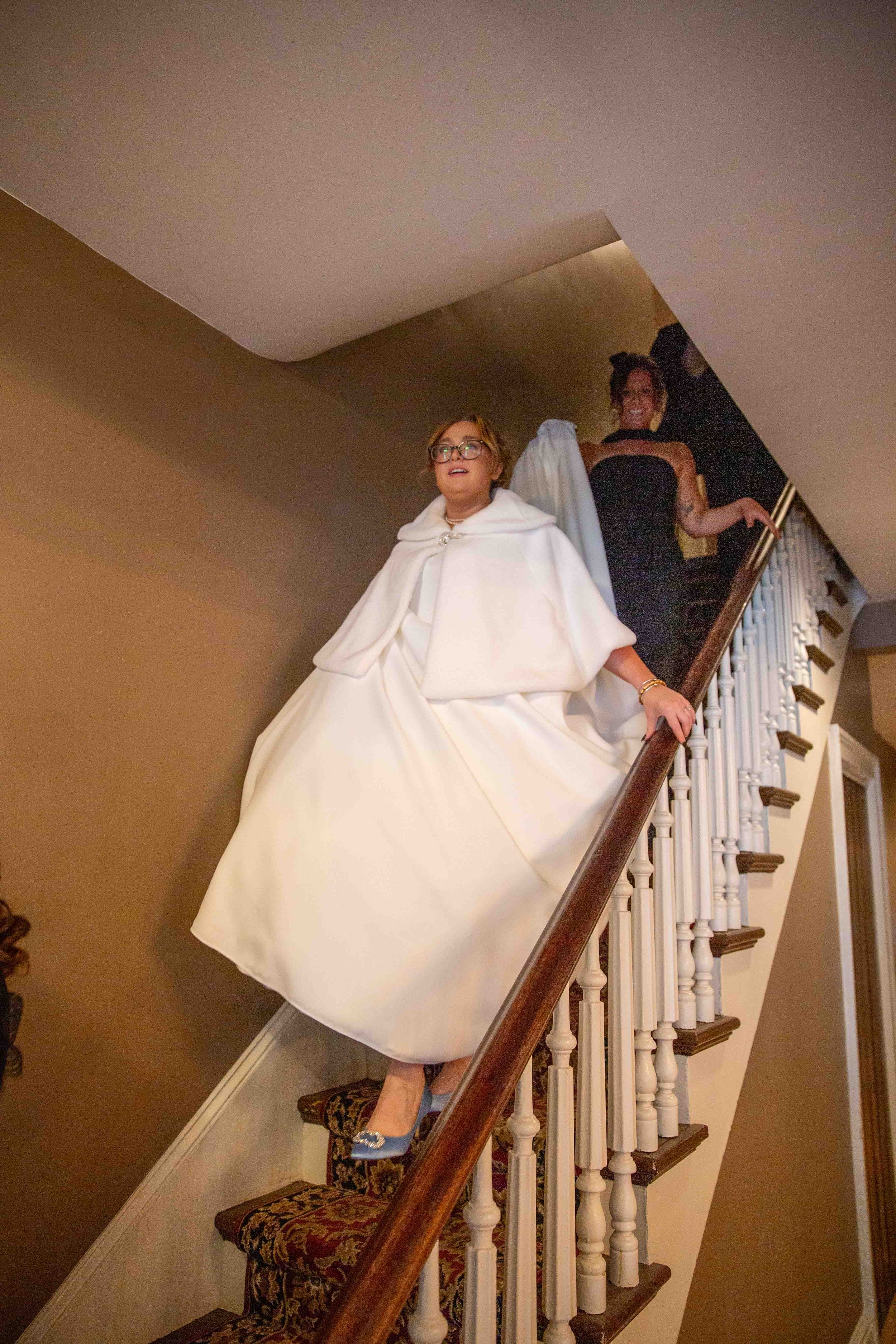 A woman in a white dress and cape descending a staircase, with another woman in a black dress at the top of the stairs.