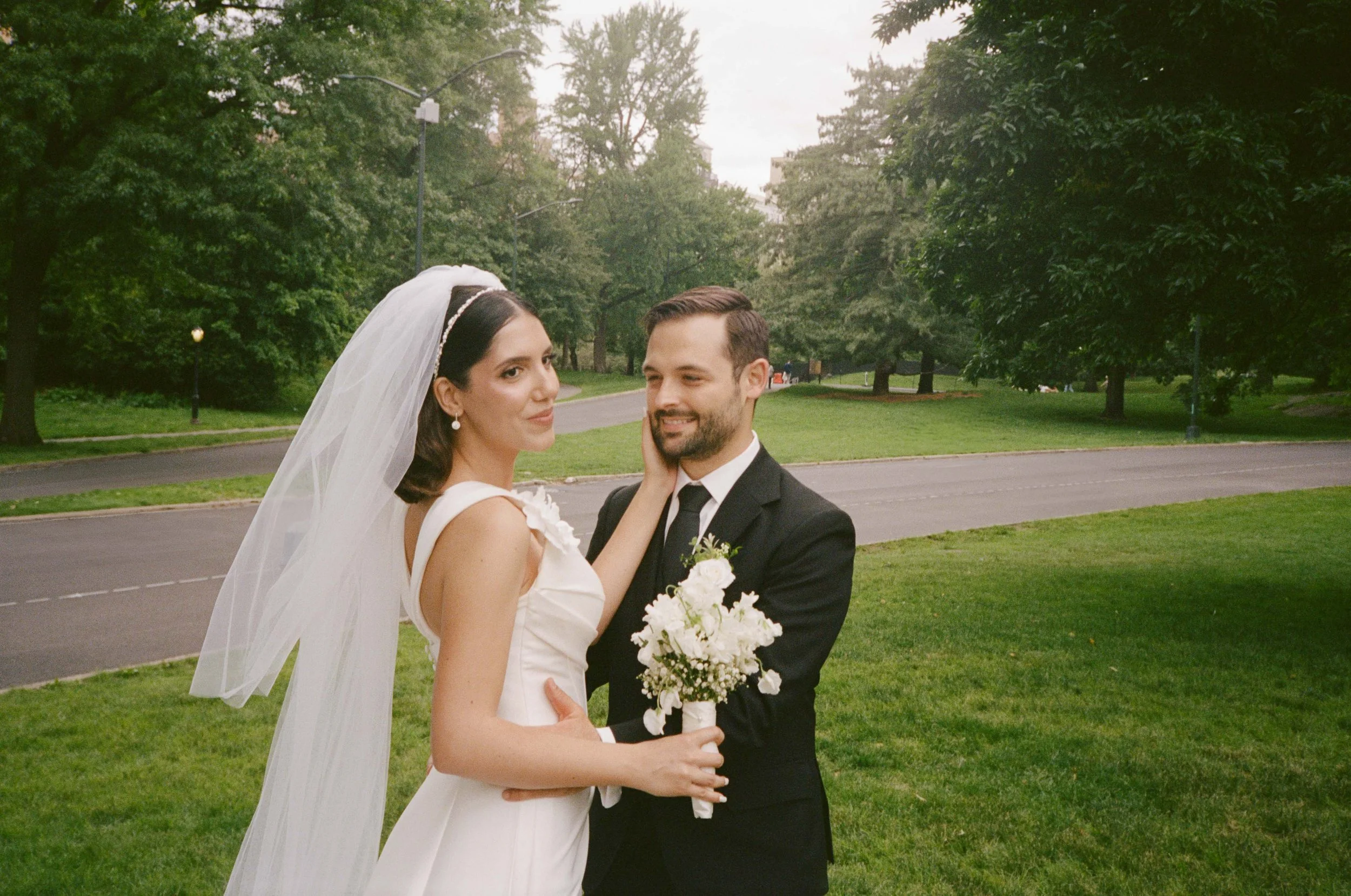 A bride and groom standing outdoors on a grassy area with trees in the background. The bride is wearing a white wedding dress with a veil and holding a bouquet of white flowers. The groom is in a black suit and tie, smiling at the bride.