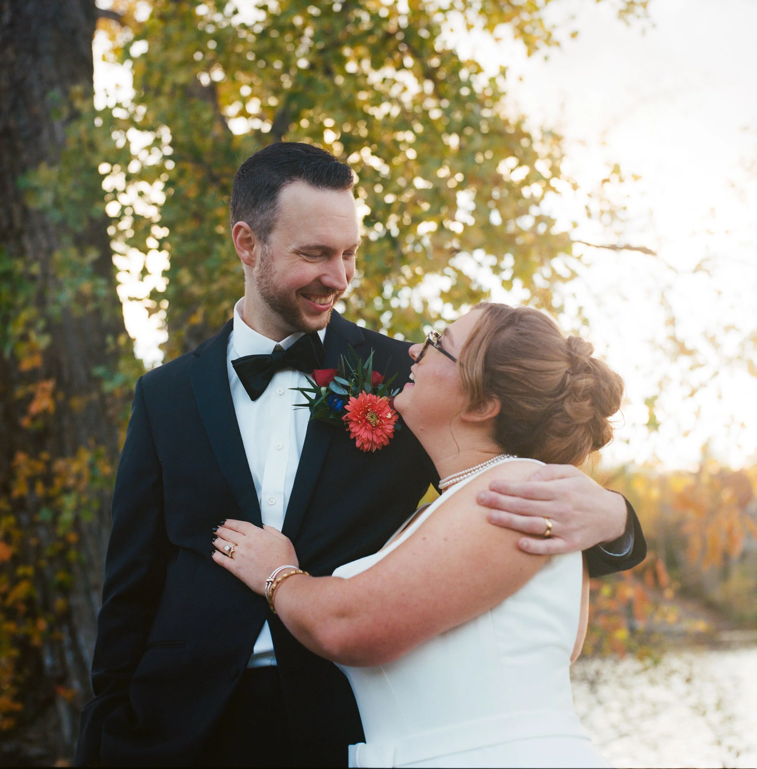 A newlywed couple embraces outdoors near a lake during sunset, with trees displaying fall foliage in the background.