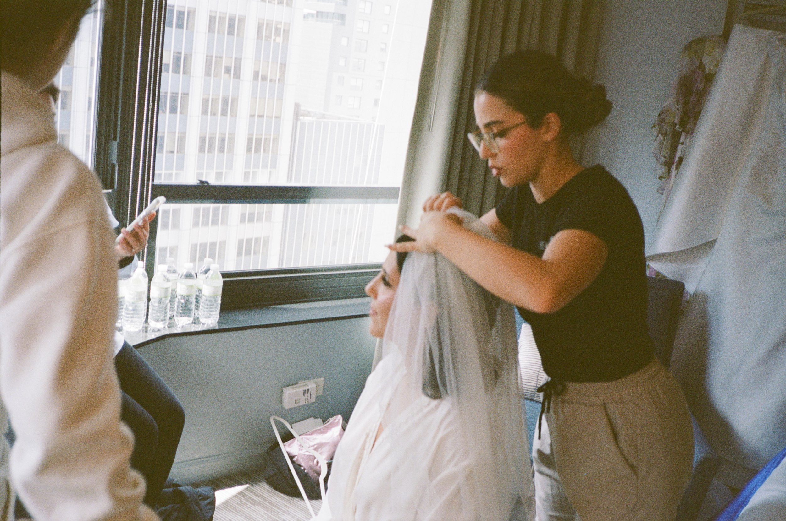 A woman is getting her hair styled by a stylist in a hotel room, with a large window showing city buildings in the background.