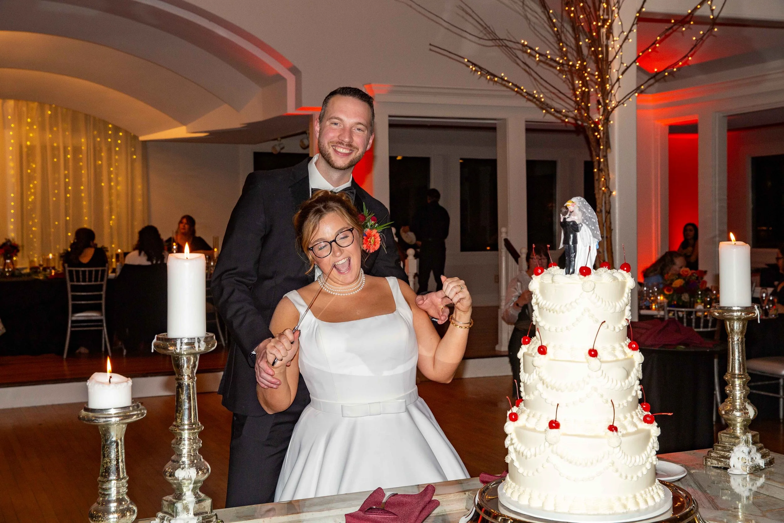 A man and woman celebrating at a wedding reception, the woman in a white dress and the man in a dark suit, standing behind a large layered wedding cake decorated with cherries and toppers of a bride and groom figurine. The woman has her eyes closed a