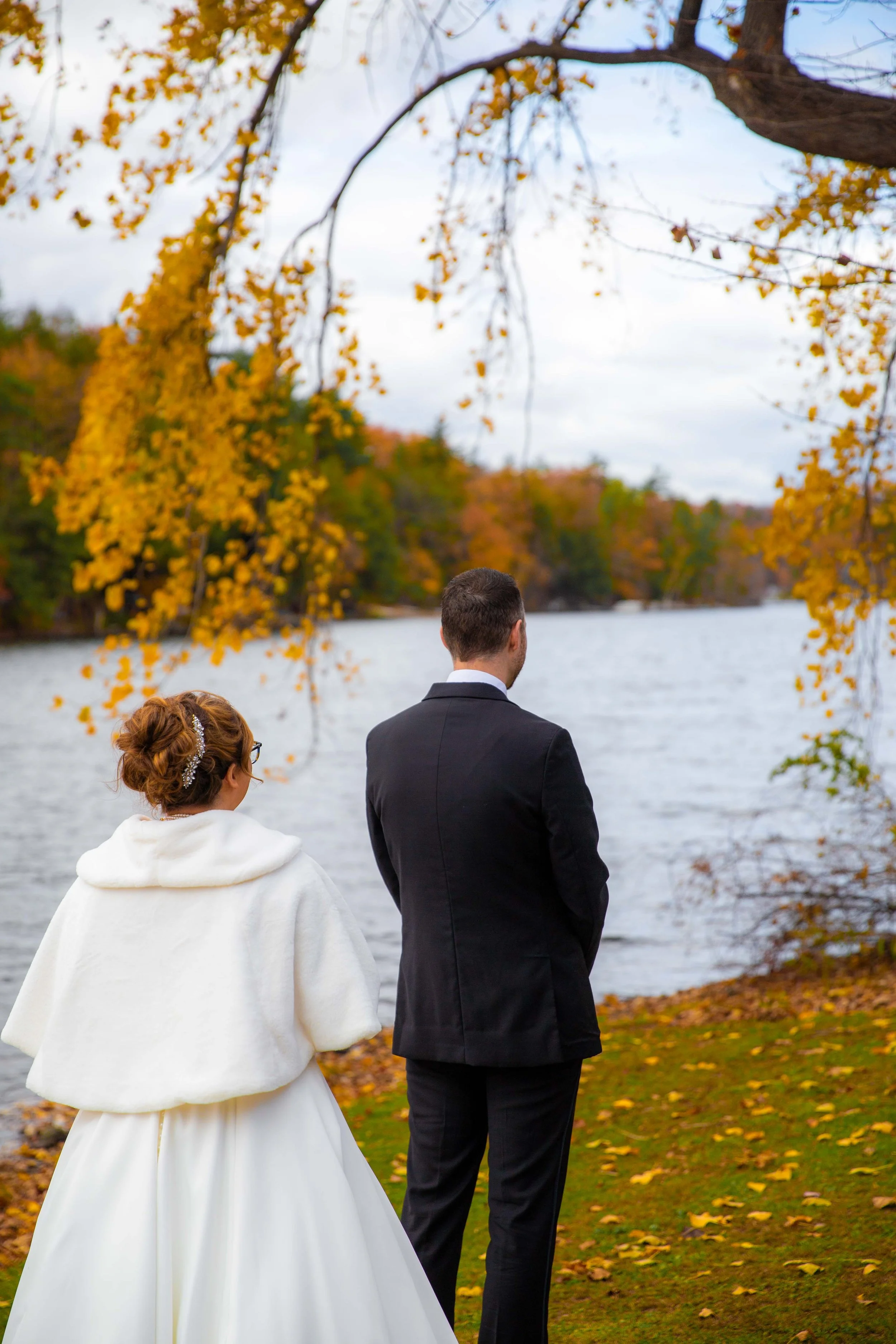 A bride and groom stand by a river during autumn, surrounded by colorful fall foliage.
