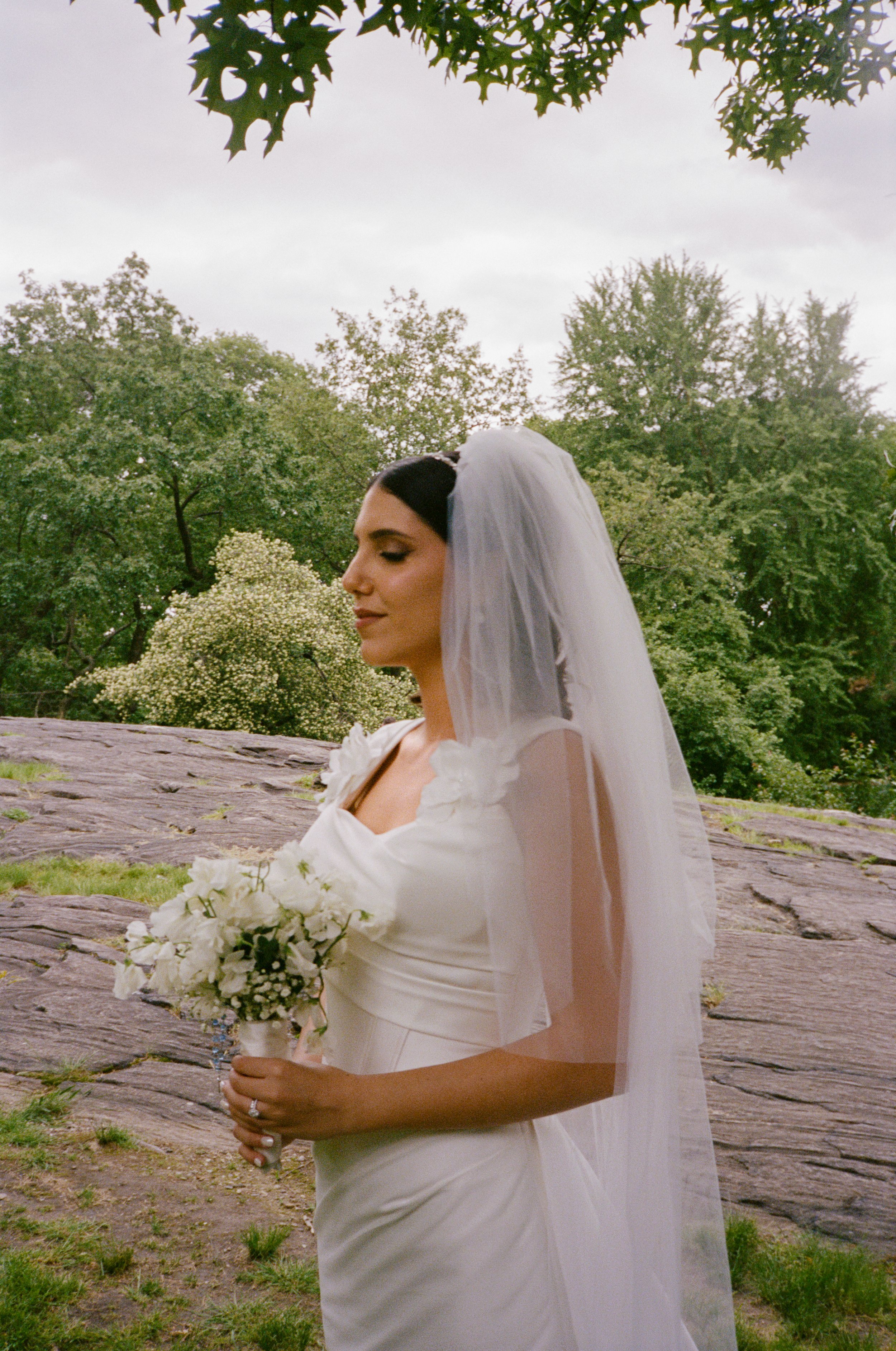 Bride in a white wedding dress with a veil, holding a bouquet of white flowers, standing outdoors on rocks with green trees in the background, and an overcast sky.