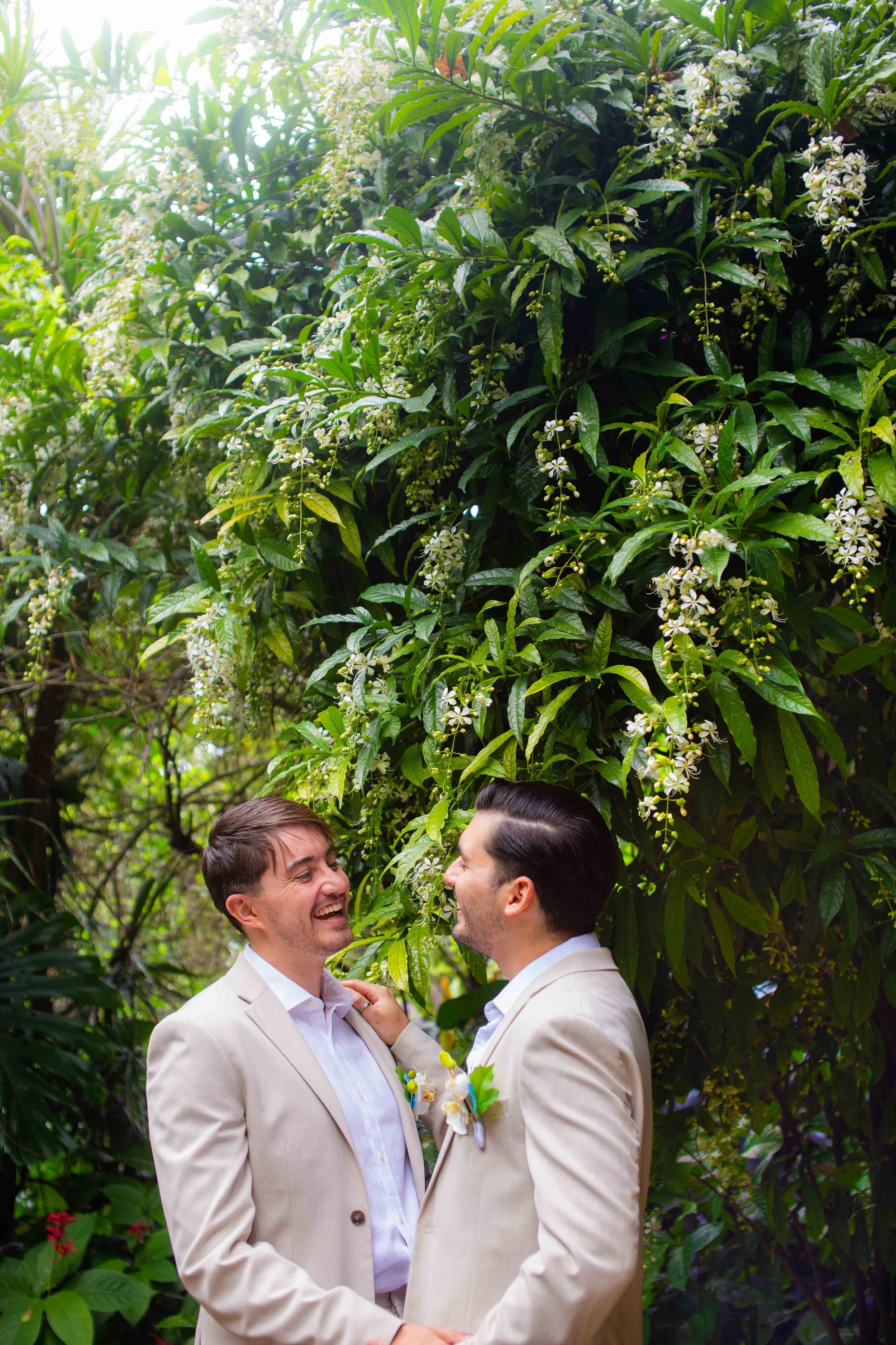 Two men in beige suits smiling and holding hands in front of lush green foliage with white flowers, celebrating a romantic moment.