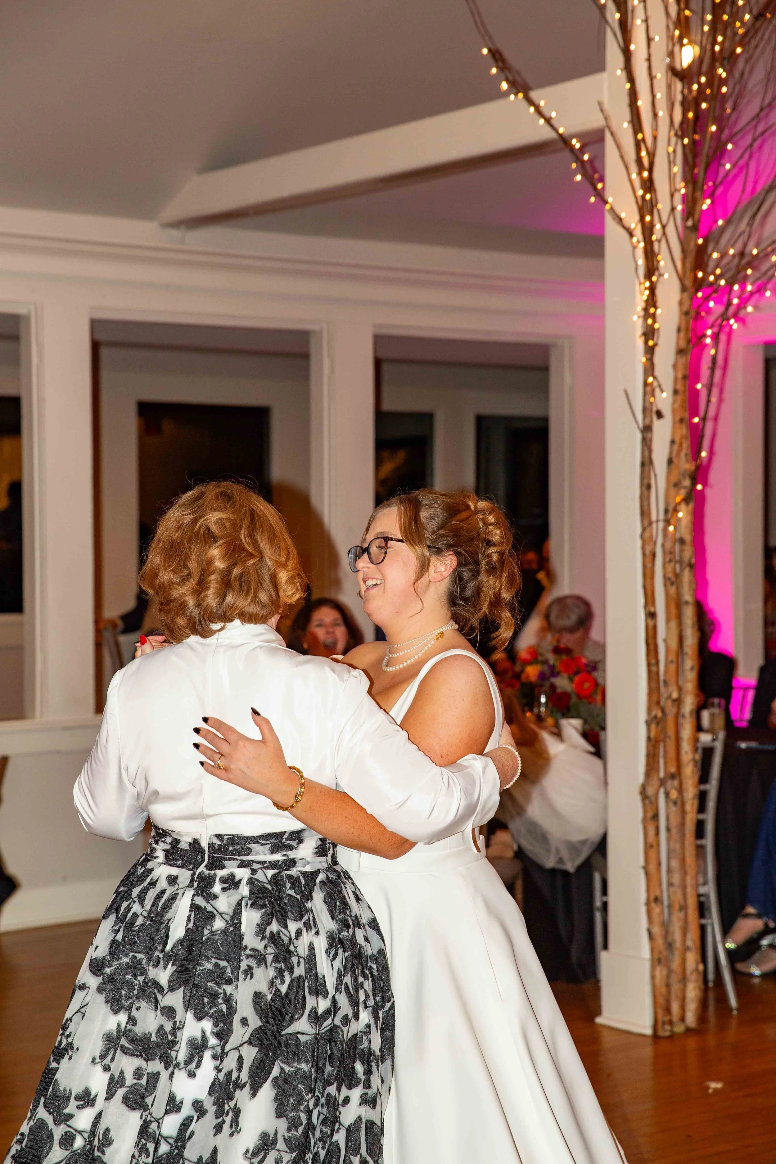 Two women are dancing and smiling at a wedding reception, one in a white wedding dress and the other in a white blouse and black-and-white floral skirt, with string lights and pink lighting in the background.