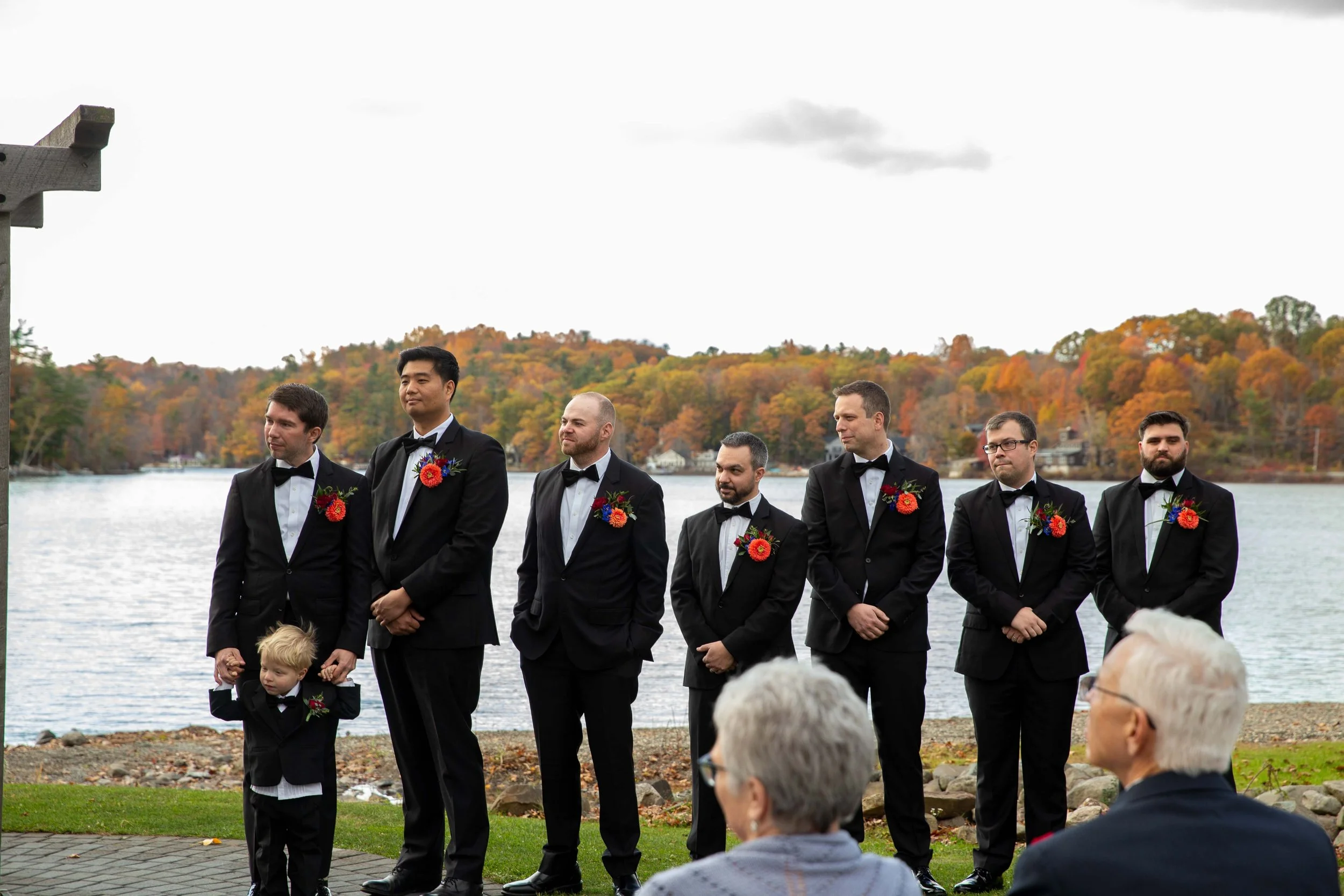 Groom and groomsmen in black tuxedos with red floral boutonnieres, standing by a lake with autumn trees, during a wedding ceremony, with an older couple in the foreground.