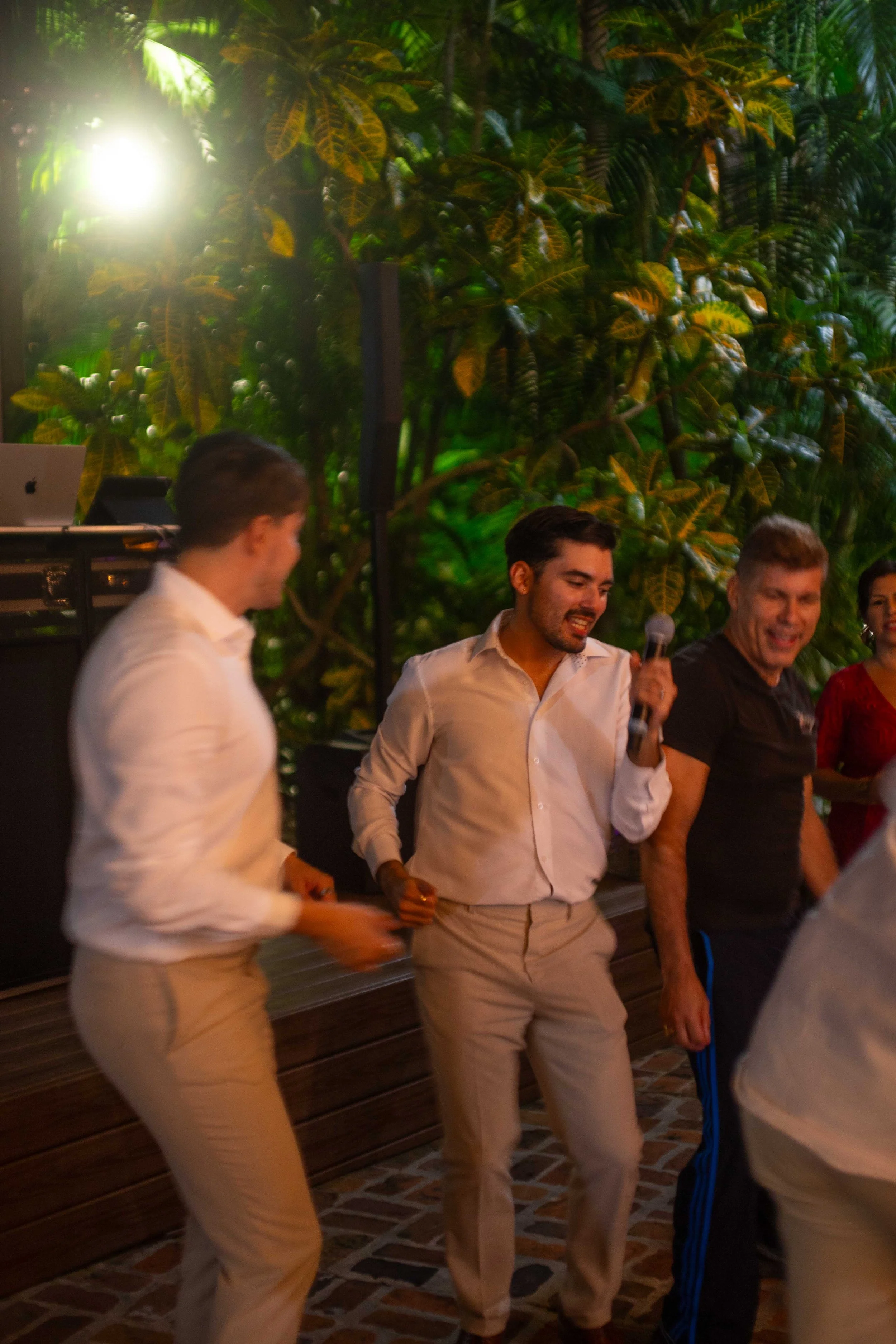 People dancing at an indoor party with lush green tropical plants in the background.