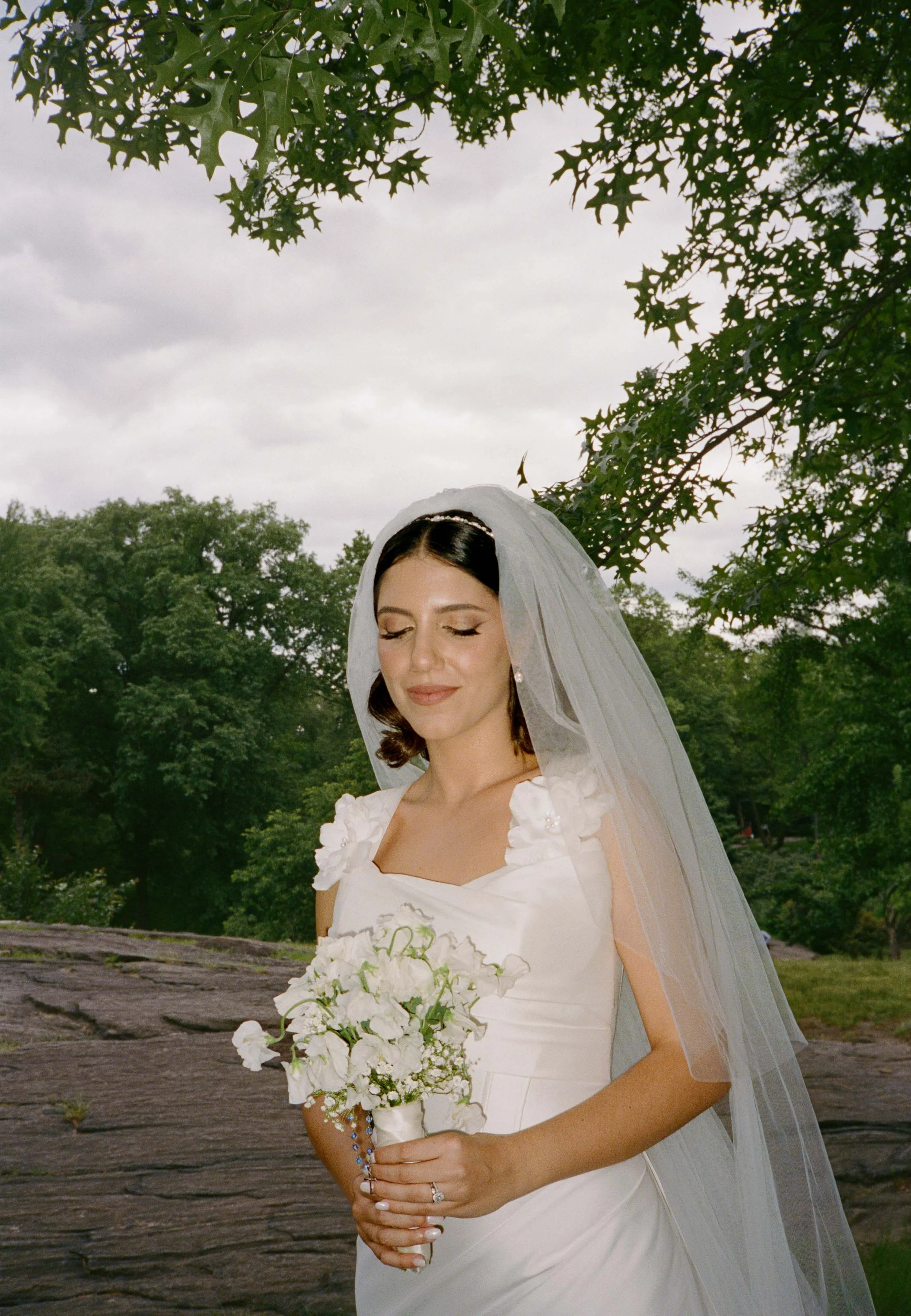A bride with dark brown hair wearing a white wedding gown with floral shoulder accents, holding a bouquet of white flowers, standing outdoors under green leafy trees with a cloudy sky in the background.