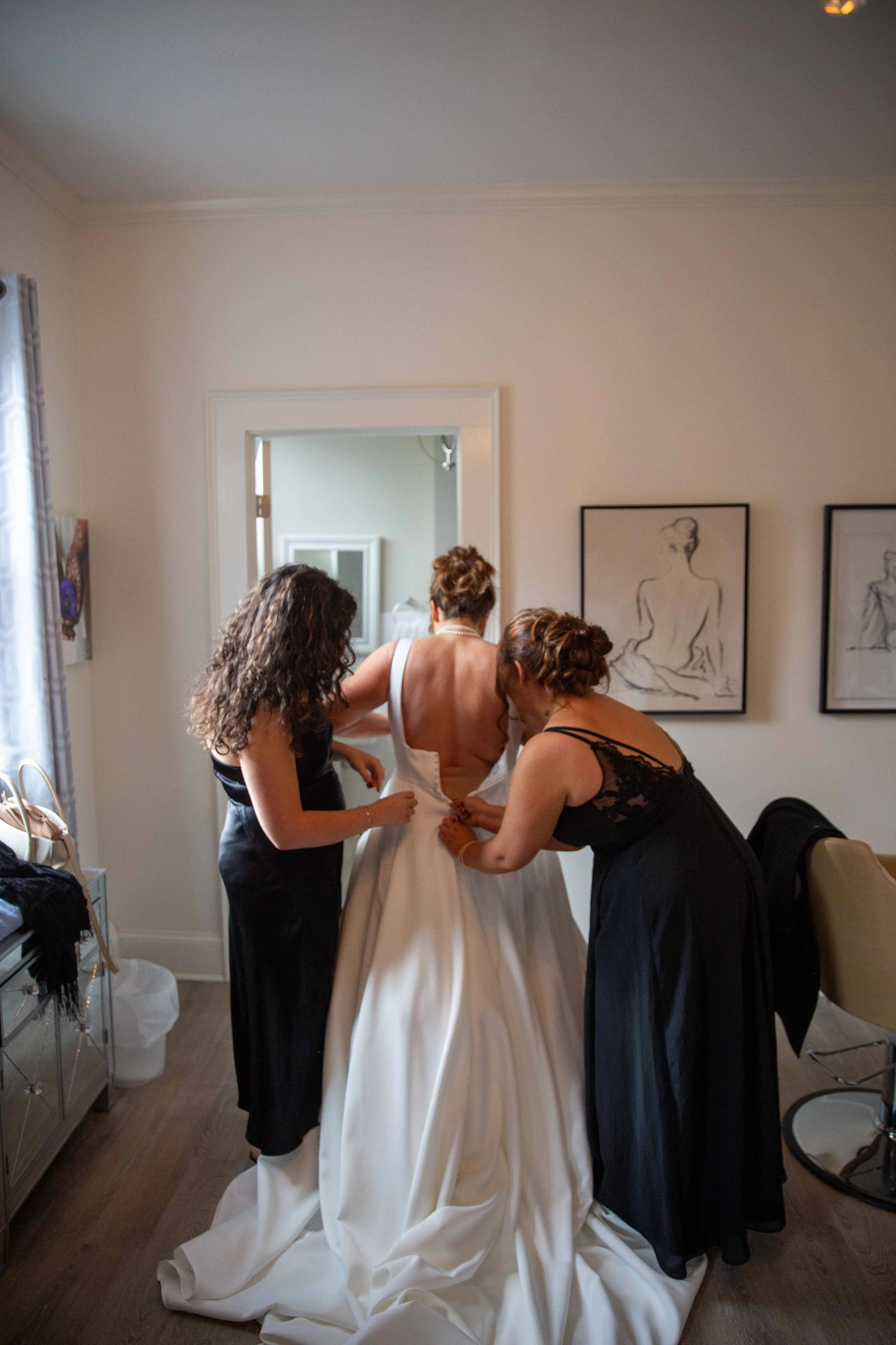 Three women in a room, one in a bridal gown being assisted by two others with final preparations for her wedding.