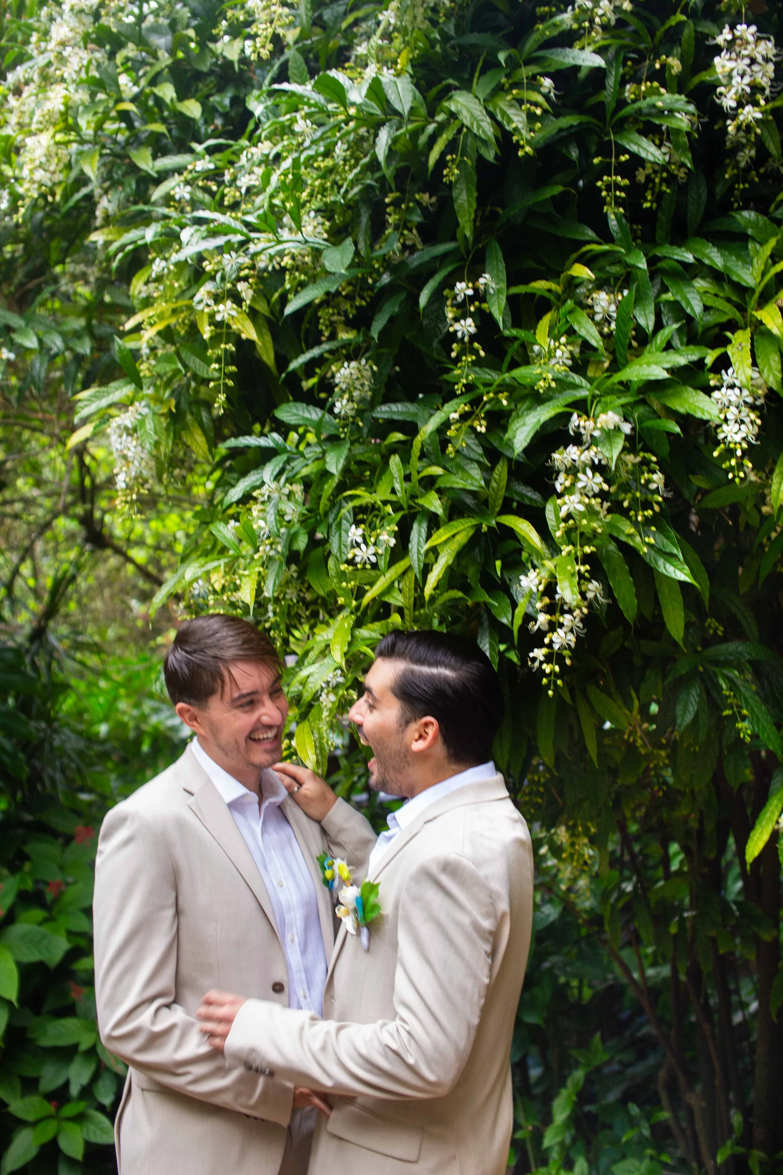 Two men in light-colored suits sharing a joyful moment outdoors in front of lush green foliage and white flowers.