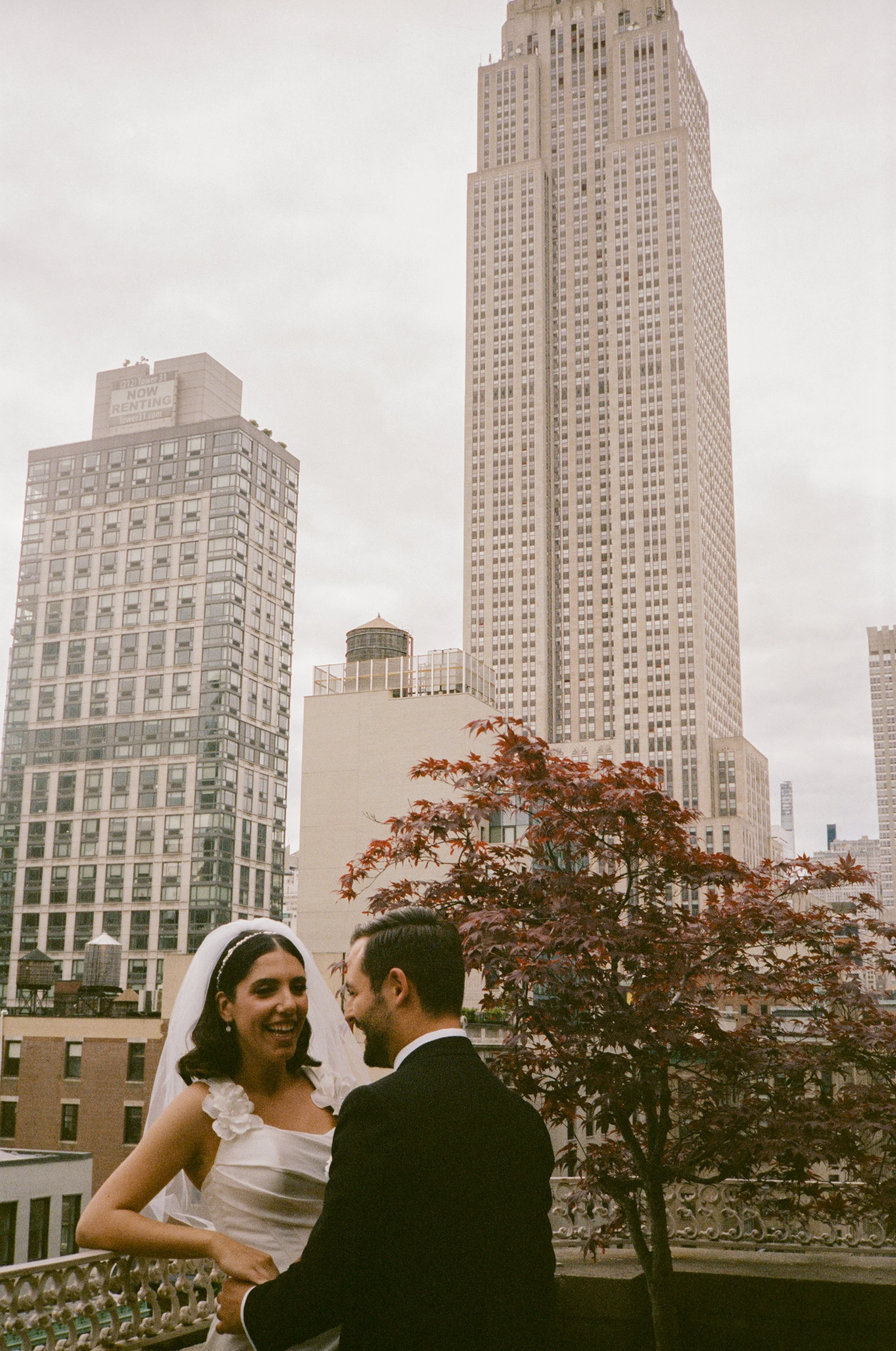 A bride and groom on a balcony with tall city buildings in the background, including the Empire State Building, during daytime.