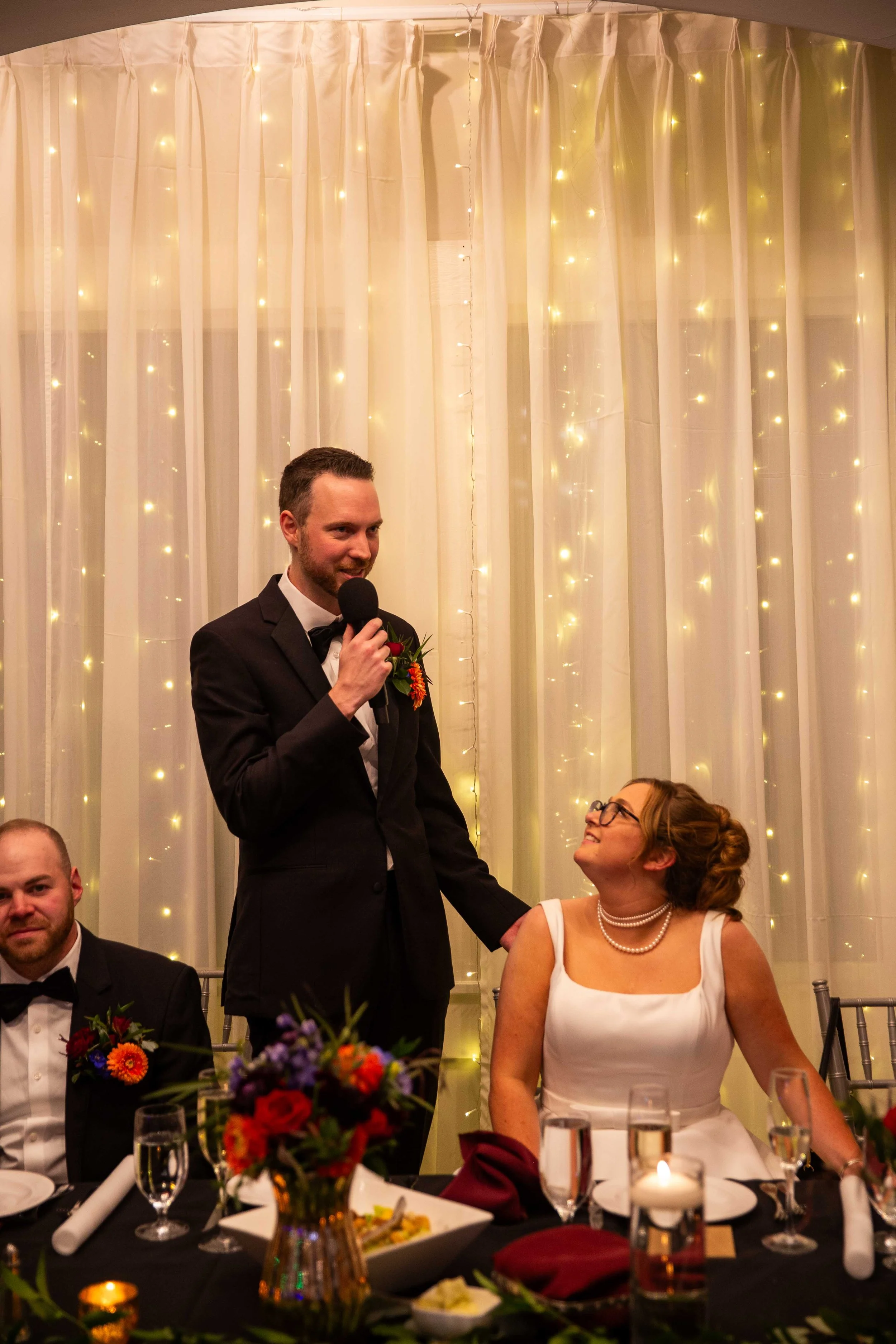 A man in a tuxedo giving a speech with a microphone while standing next to a bride seated at a wedding reception table. The bride is wearing a white dress, pearl necklace, and glasses, looking up at the man. The background features white curtains wit