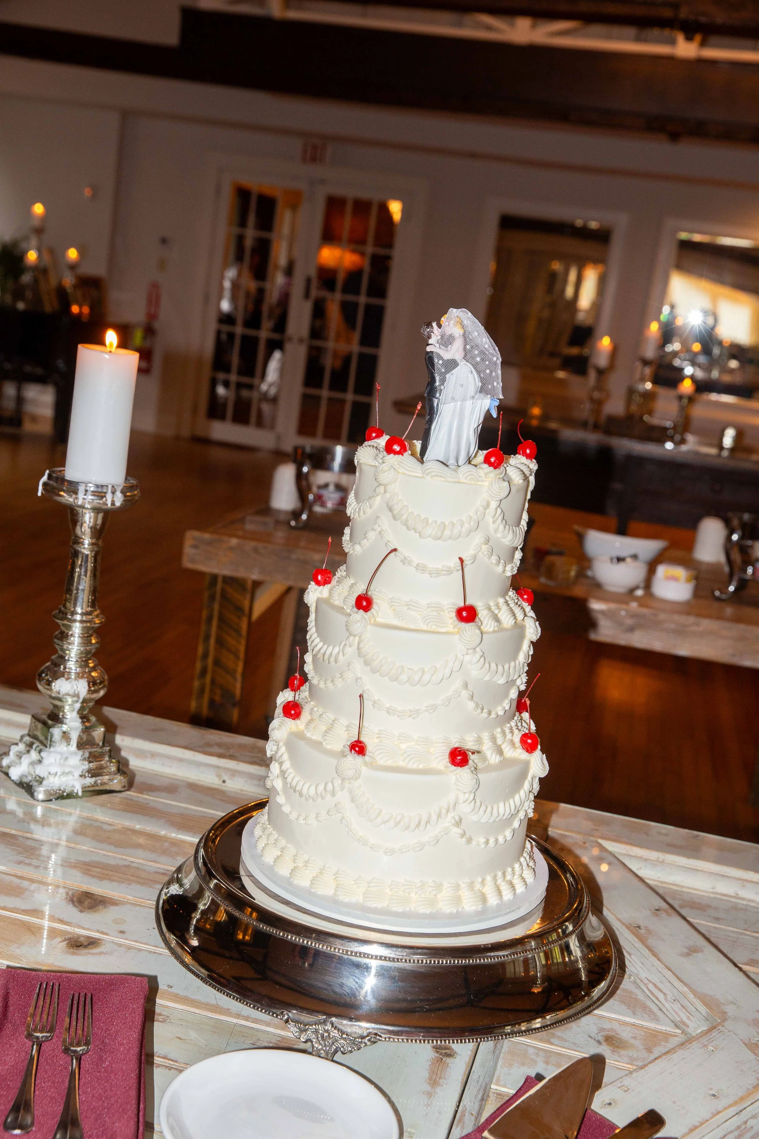 Four-tiered wedding cake with white frosting, decorated with cherries and pearl-like icing. Cake topper features a figurine of a bride and groom.